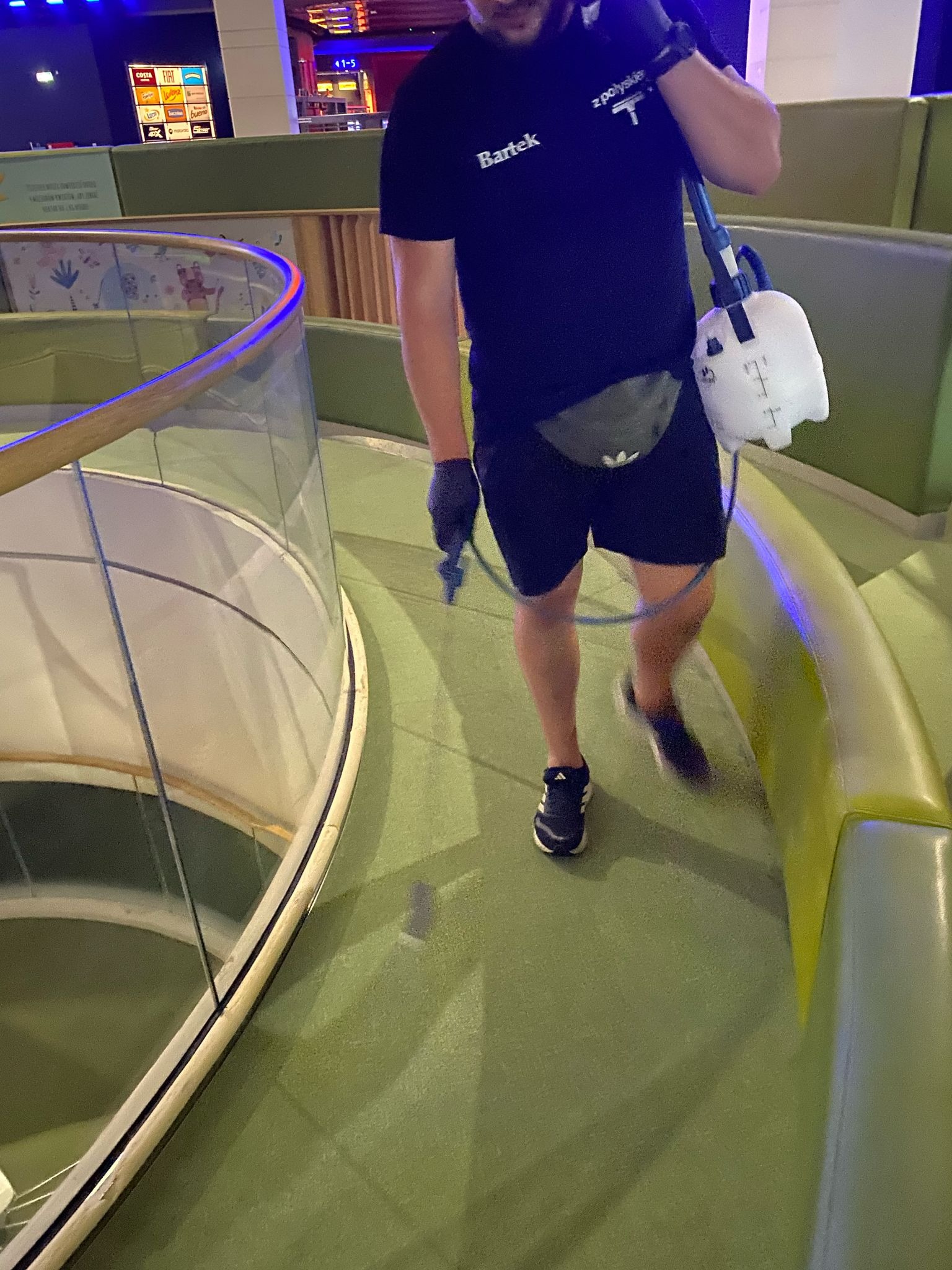 Pracownik w Łukowie dezynfekuje podłogę w centrum handlowym, używając opryskiwacza. Widoczna część balustrady i oznaczenia sklepów w tle.