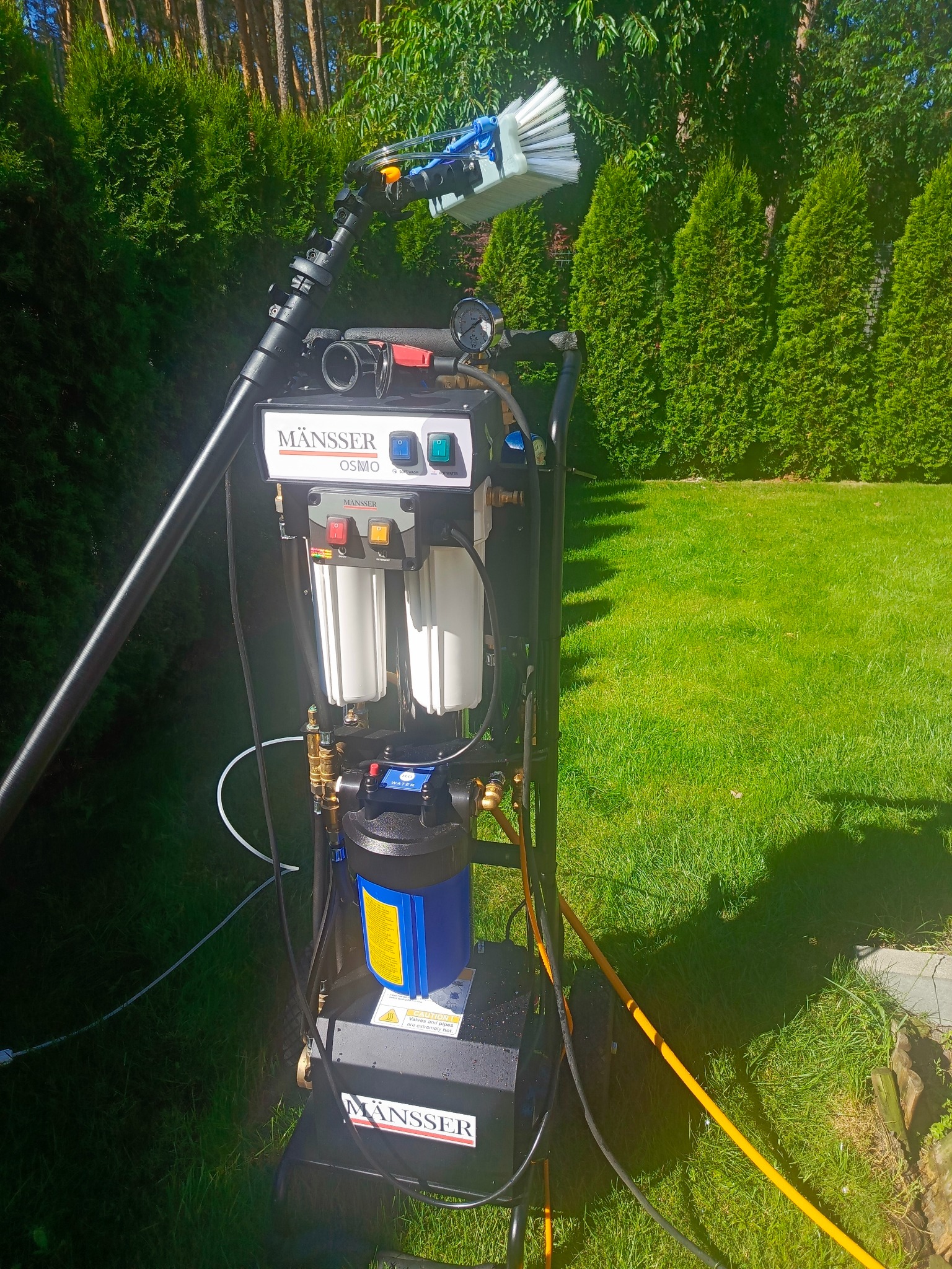 Profesjonalny sprzęt do czyszczenia MÄNSSER OSMA na trawie, z wysuwaną szczotką i filtrami, gotowy do pracy w ogrodzie. Tuje w tle.