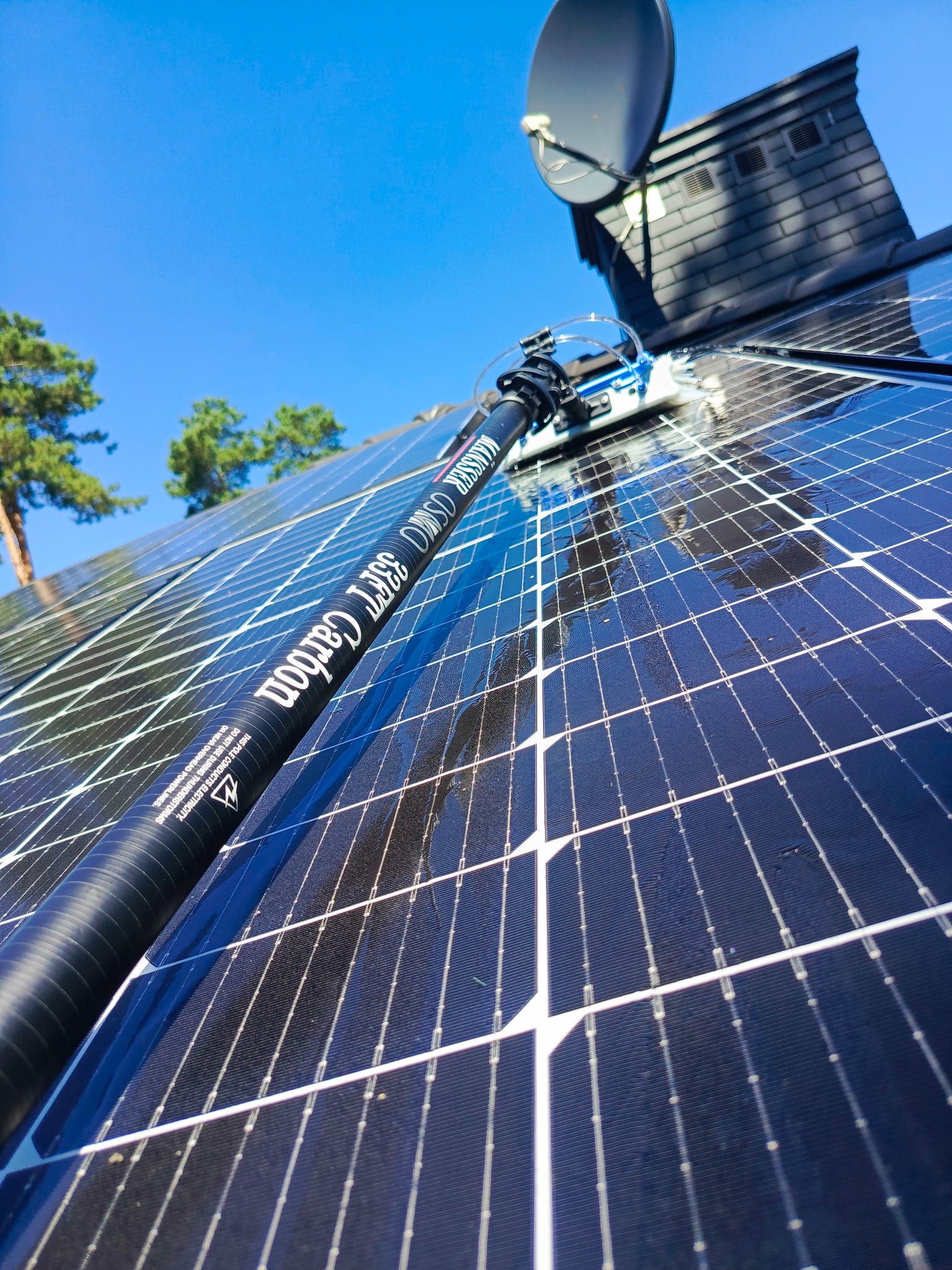 Czyszczenie paneli słonecznych długą lancą teleskopową z szczotką, w tle dach budynku z anteną satelitarną i fragmenty drzew na tle błękitnego nieba.
