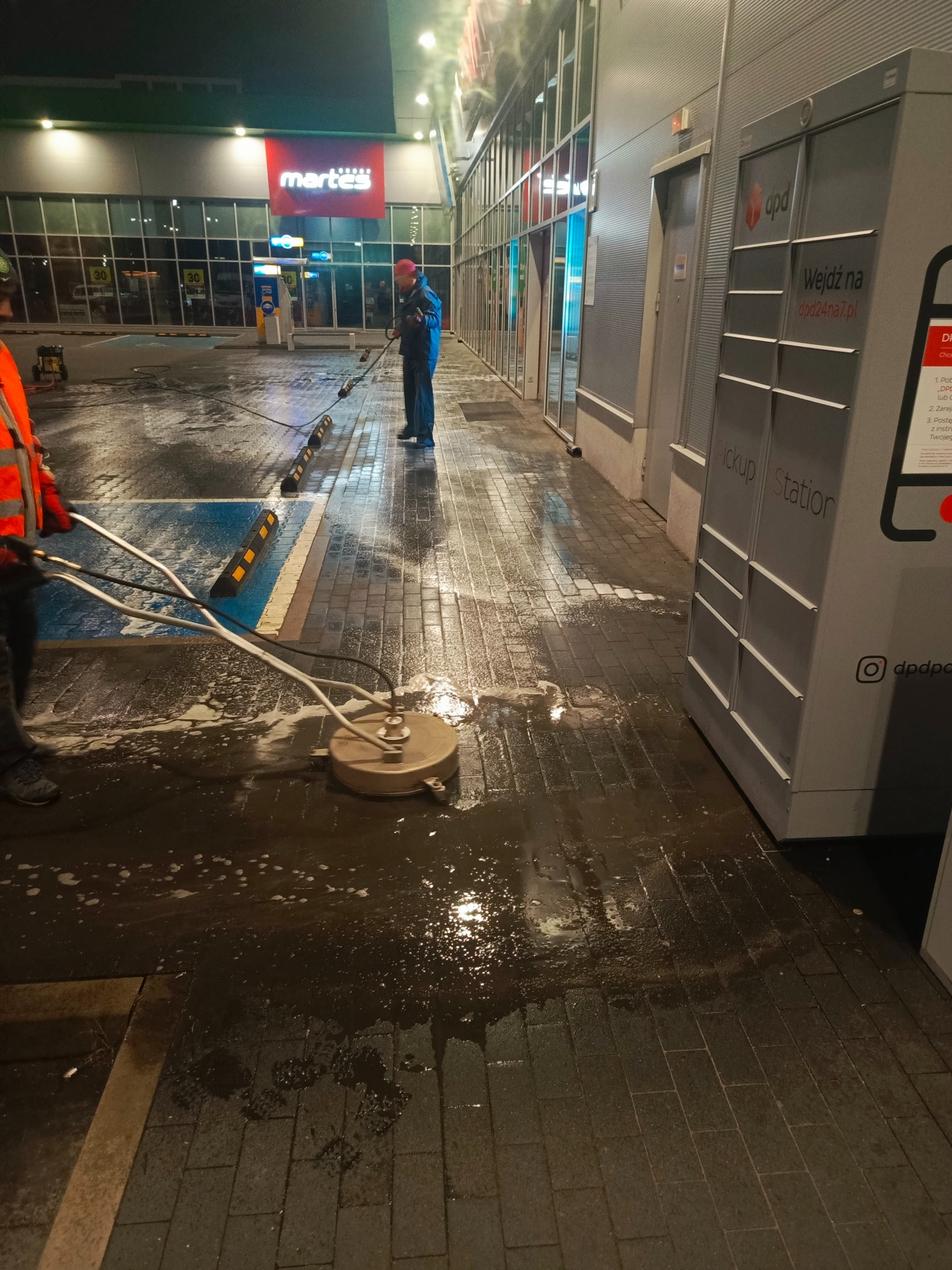Dwóch mężczyzn czyści kostkę brukową przed budynkiem sklepu Martex i automatem DPD. Jeden używa specjalnej maszyny, drugi lancy ciśnieniowej, widać mokrą powierzchnię.