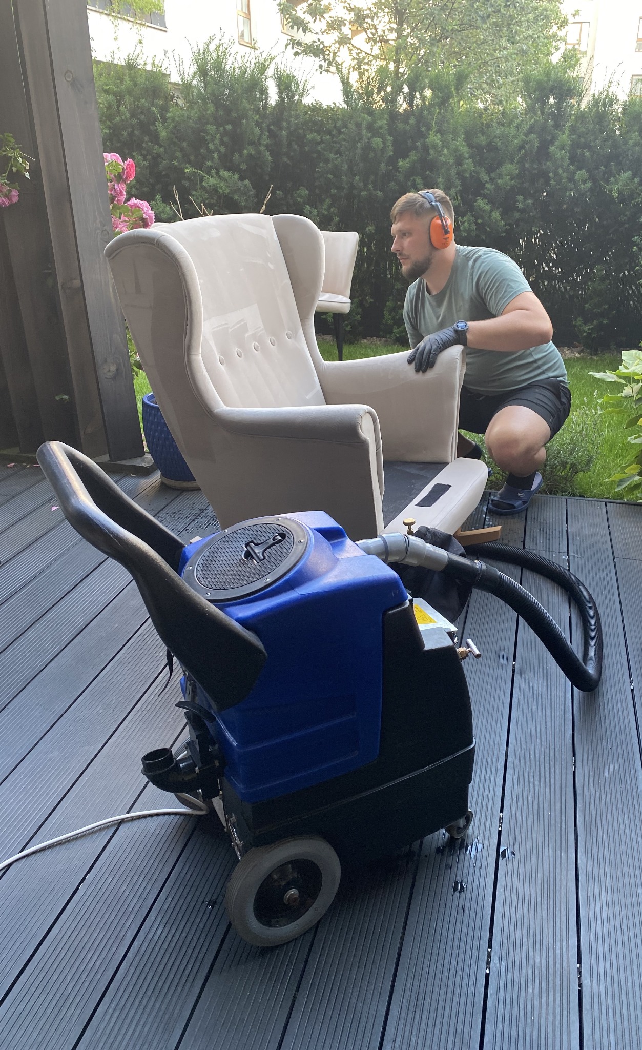 Pracownik w słuchawkach przygotowuje się do czyszczenia beżowego fotela za pomocą profesjonalnego sprzętu na drewnianym tarasie z zielonym tłem.