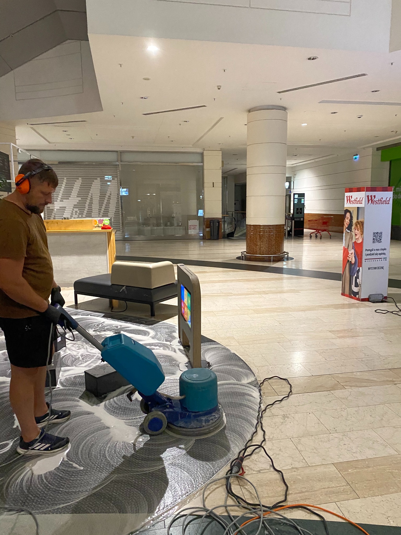 Pracownik w słuchawkach czyści maszynowo wzorzystą podłogę w centrum handlowym Westfield. Widoczny automat czyszczący i kabel zasilający.