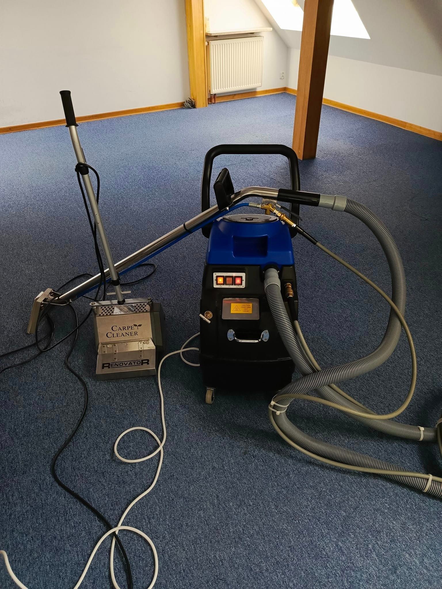 Profesjonalny sprzęt do czyszczenia dywanów marki Renovator na tle niebieskiej wykładziny w jasnym pomieszczeniu. Widoczne węże i elementy robocze urządzenia.