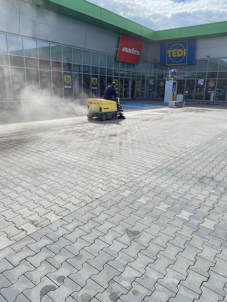 Operator na maszynie Kärcher czyści kostkę brukową przed sklepem Martes i Tedi. Widoczna para wodna unosząca się znad czyszczonej powierzchni.