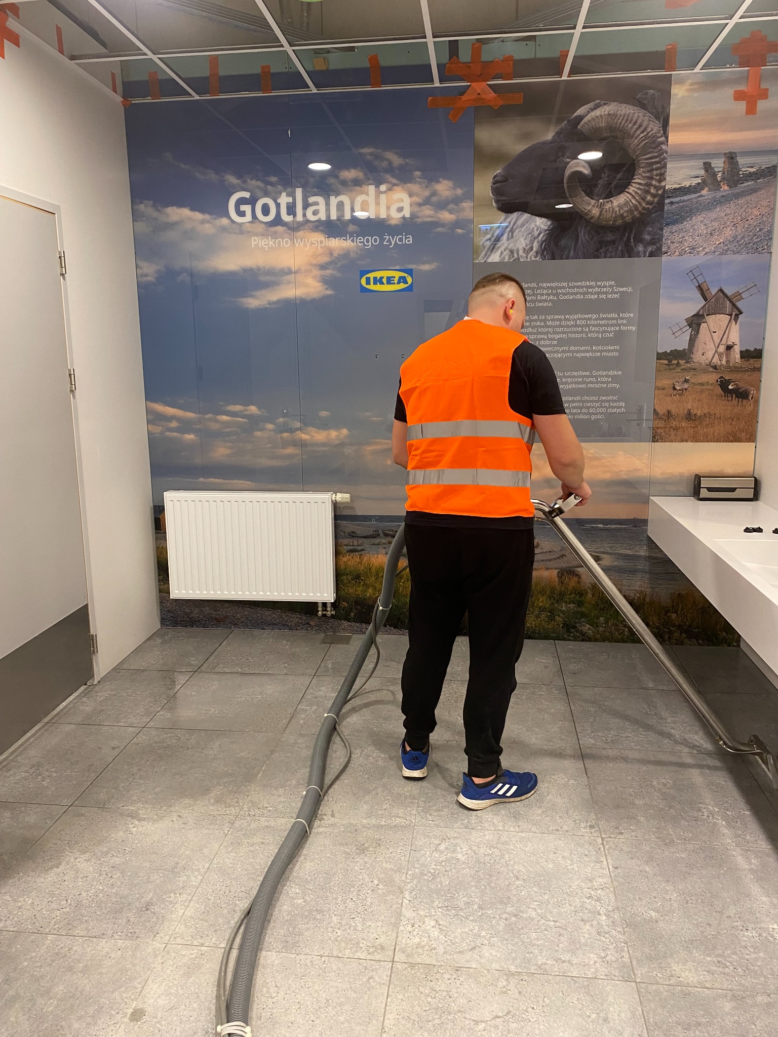 Pracownik w kamizelce czyści podłogę w pomieszczeniu z grafiką Gotlandii i logo IKEA, używając profesjonalnego sprzętu do czyszczenia podłóg.