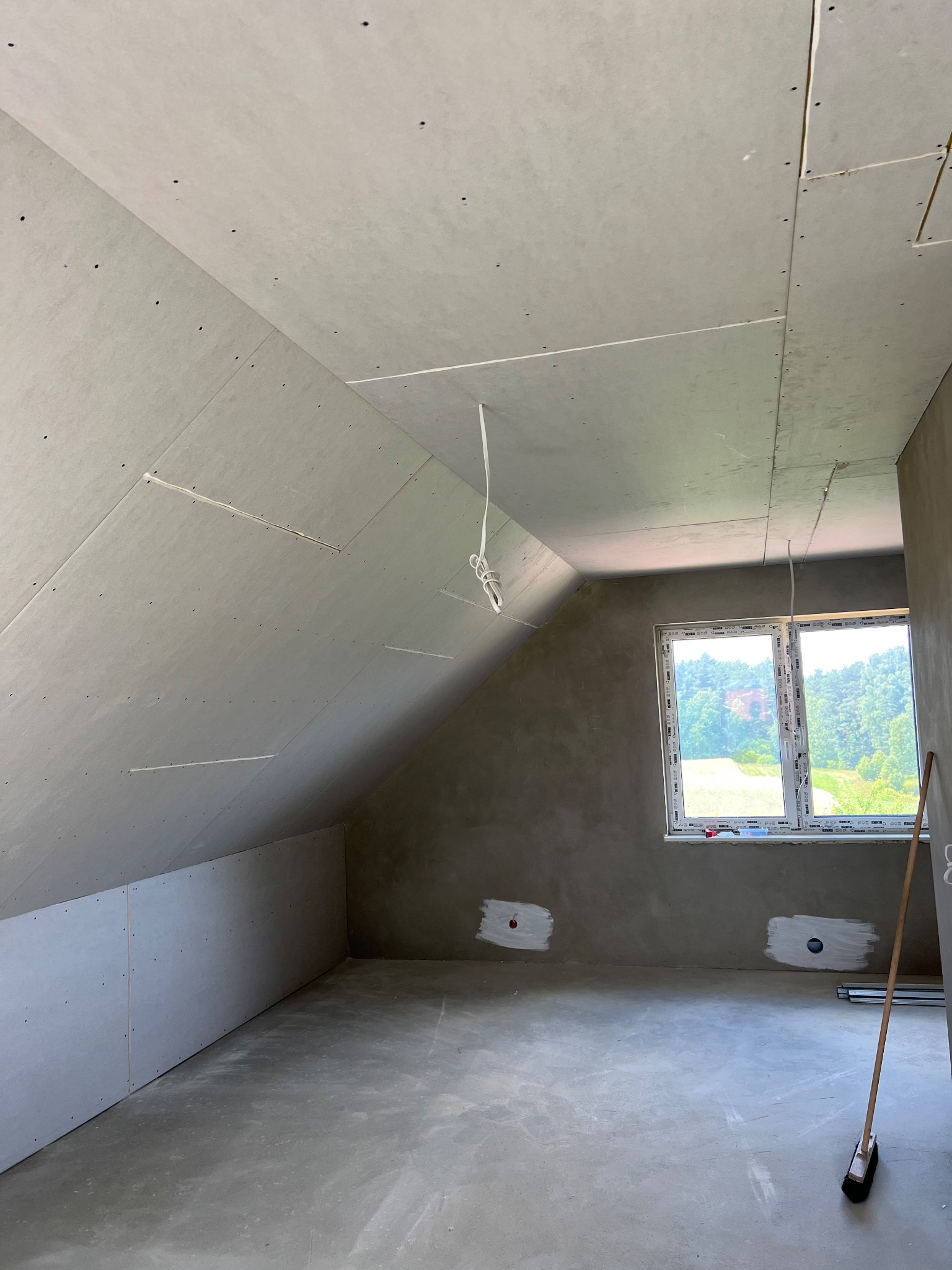 Poddasze z oknem w trakcie montażu płyt kartonowo-gipsowych na skosach sufitu i ścianach, widoczne wystające przewody elektryczne i przygotowane otwory pod gniazdka.