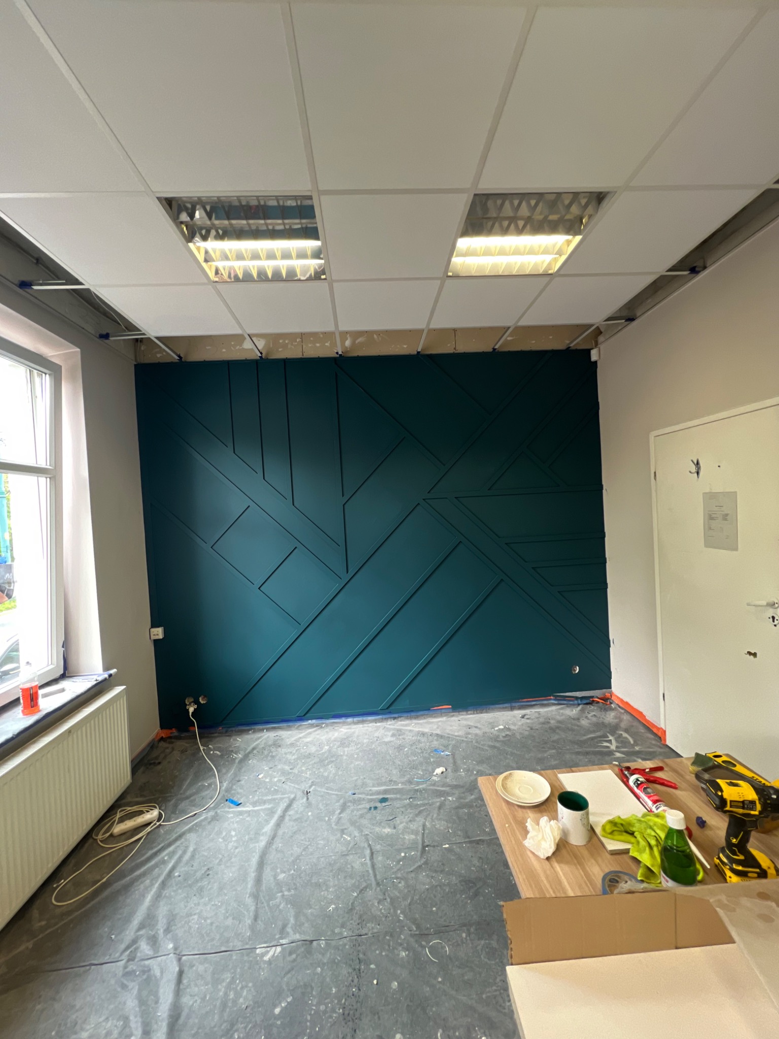 Montaż dekoracyjnej, geometrycznej ściany z paneli w kolorze morskim, widoczne narzędzia i zabezpieczona folią podłoga, sufit kasetonowy z lampami rastrowymi.