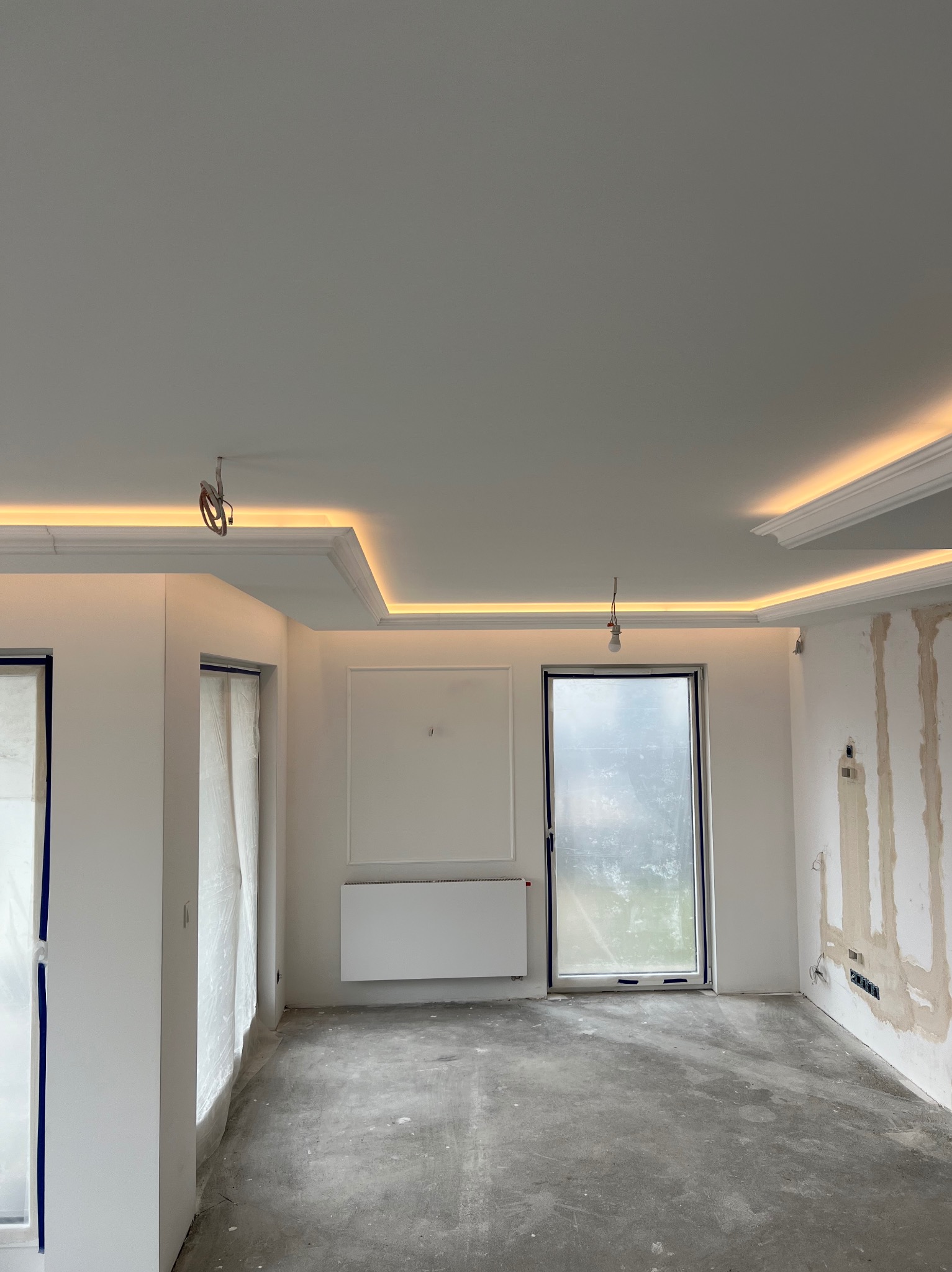 Pokój w trakcie prac wykończeniowych: surowa podłoga, ściany częściowo przygotowane do malowania, ozdobne listwy sufitowe z podświetleniem LED, okna zabezpieczone folią.