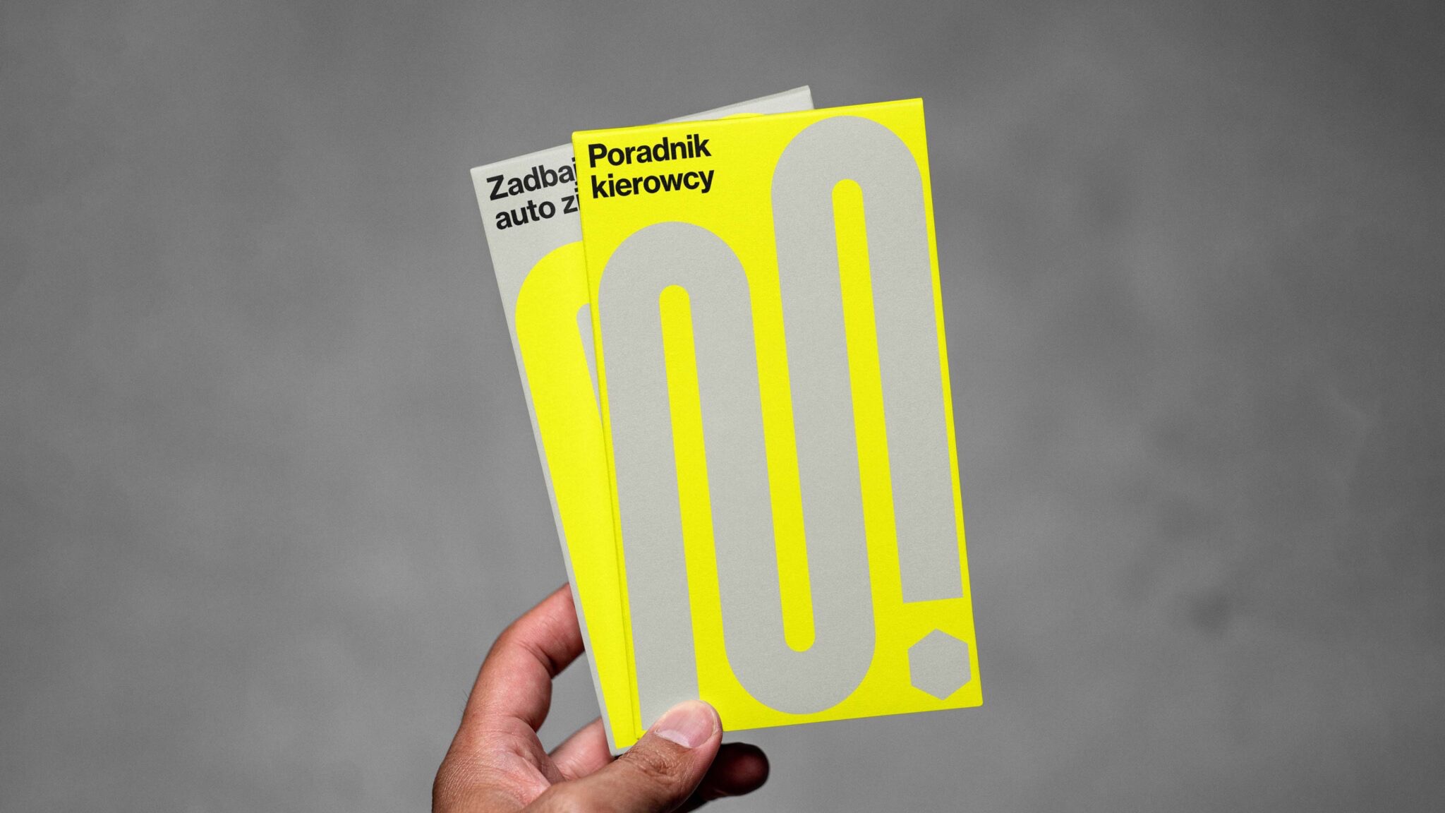 Dwa poradniki dla kierowców w odcieniach szarości i żółci, trzymane w dłoni, z minimalistycznym wzorem i typografią.