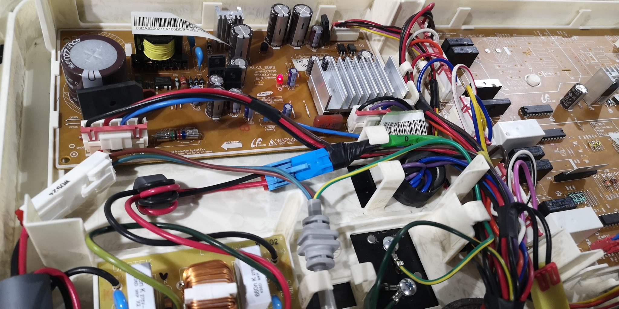 Wnętrze obudowy urządzenia AGD z widoczną płytą główną, kondensatorami, przewodami w różnych kolorach i złączami, z bliska.