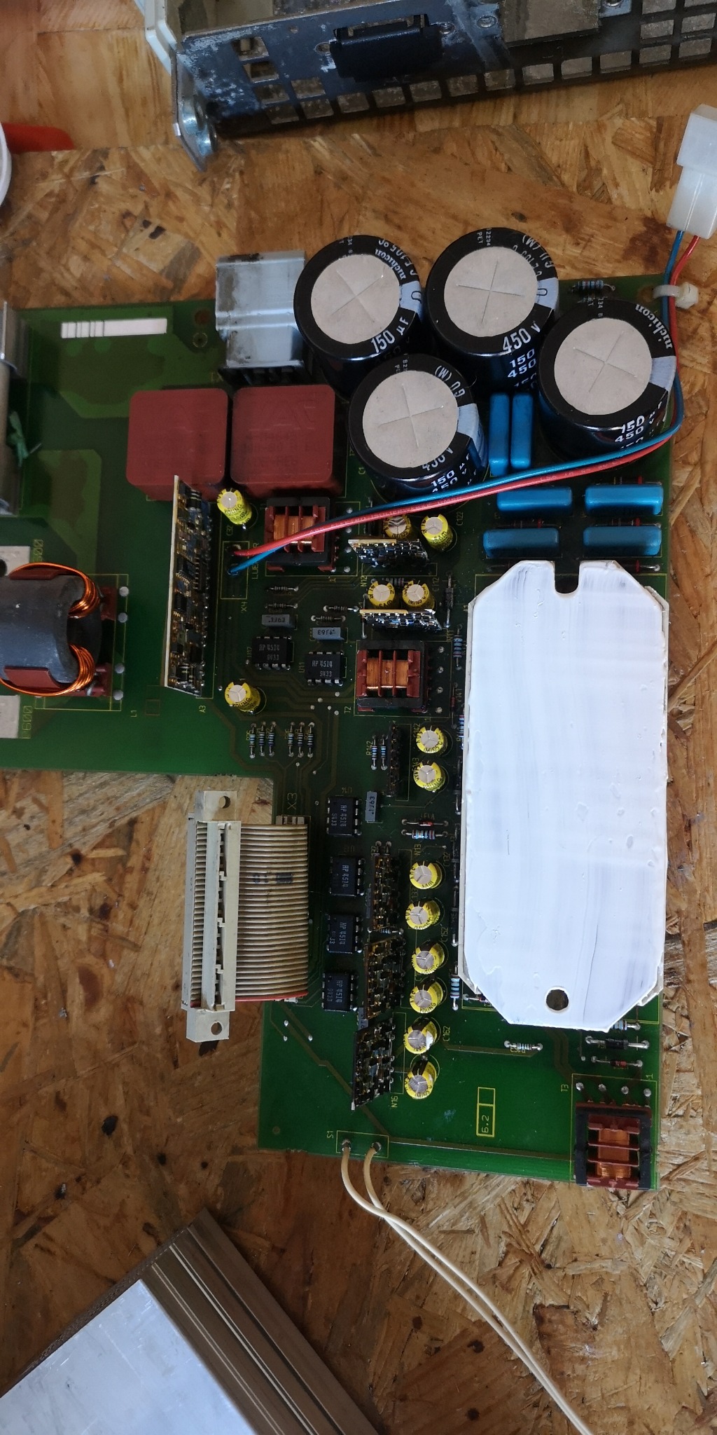 Zielona płyta elektroniczna z kondensatorami, transformatorami i innymi elementami, widoczna z góry na tle drewnianej powierzchni, prawdopodobnie podczas naprawy urządzenia AGD.