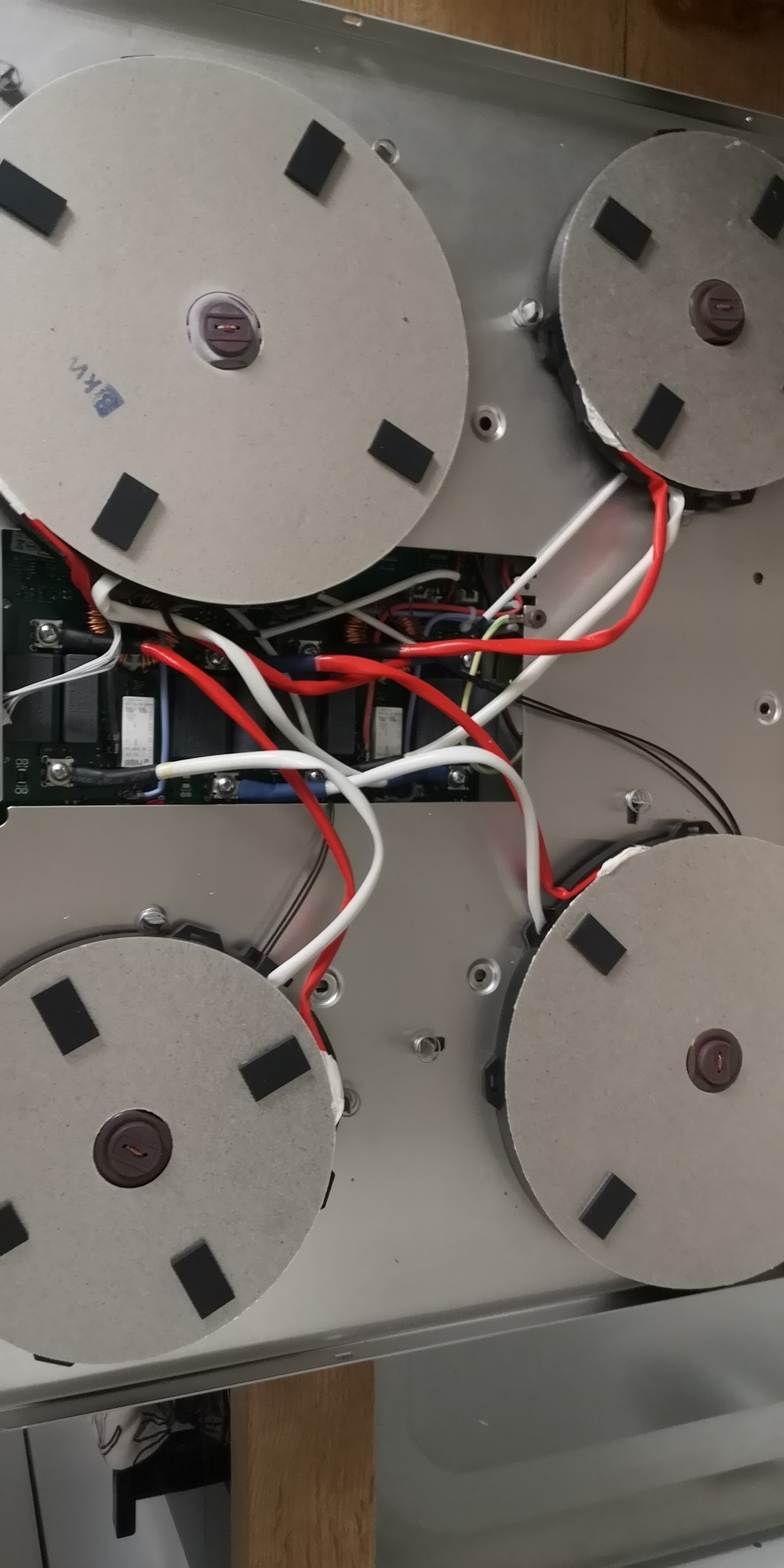 Wnętrze płyty indukcyjnej z widocznymi cewkami, przewodami w izolacji czerwonej i białej oraz elementami elektroniki na płytce drukowanej.