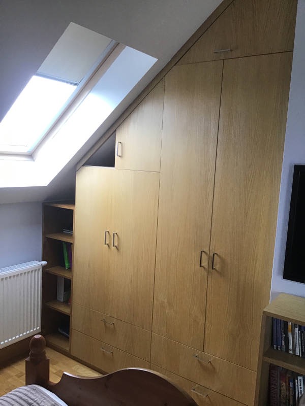 Zabudowa szafy wnękowej z drewna w pokoju na poddaszu ze skosem i oknem dachowym, dopasowana do kształtu dachu.