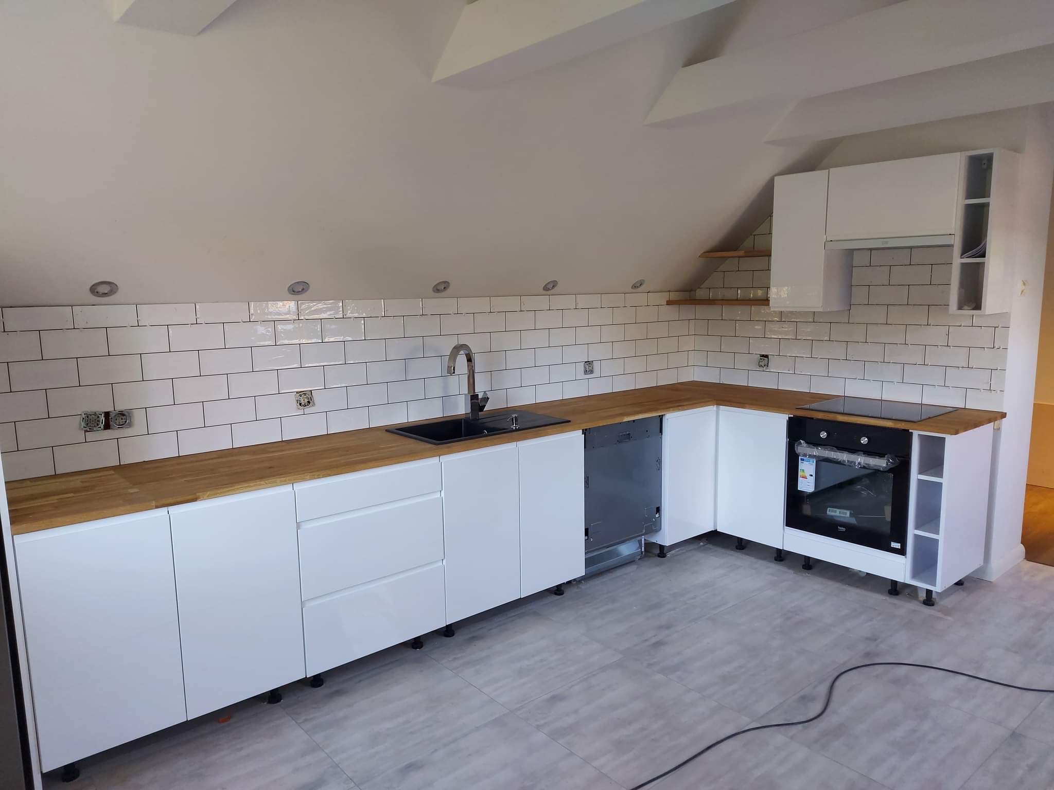 Kuchnia w trakcie remontu w skosach: białe szafki z drewnianym blatem, białe płytki na ścianie, widoczne gniazdka elektryczne przed montażem osprzętu, szare płytki podłogowe.