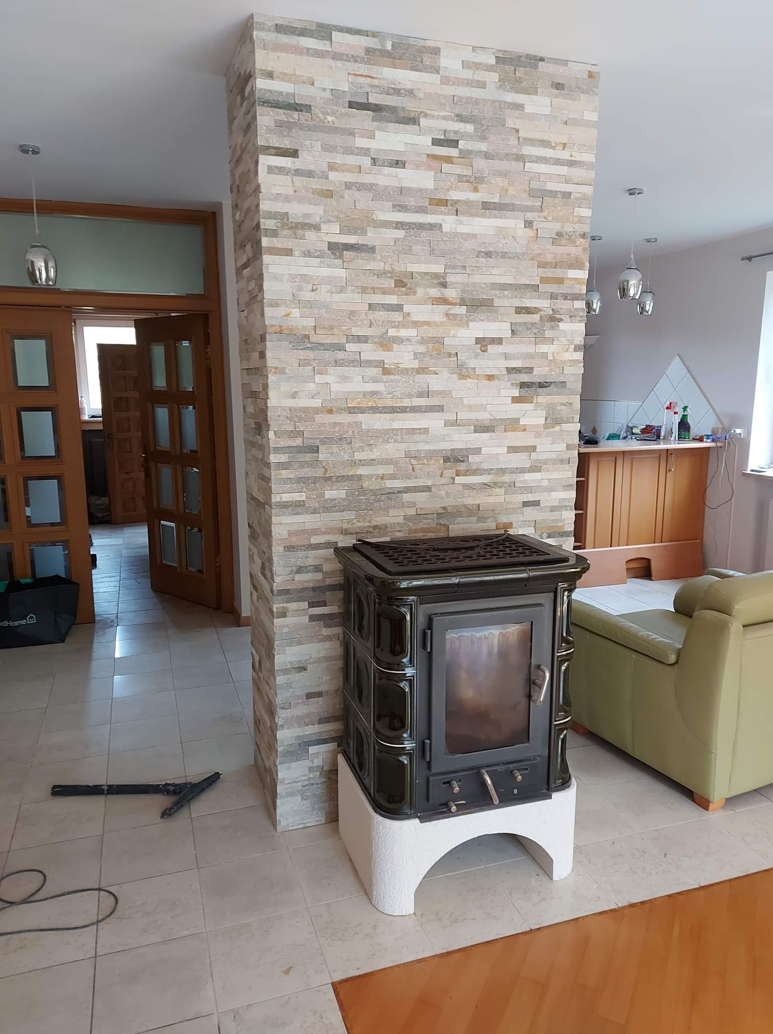 Wnętrze z kolumną obłożoną dekoracyjnym kamieniem, na której stoi czarny piec kaflowy na białej podstawie, widoczny fragment jasnej sofy i podłoga wyłożona płytkami.