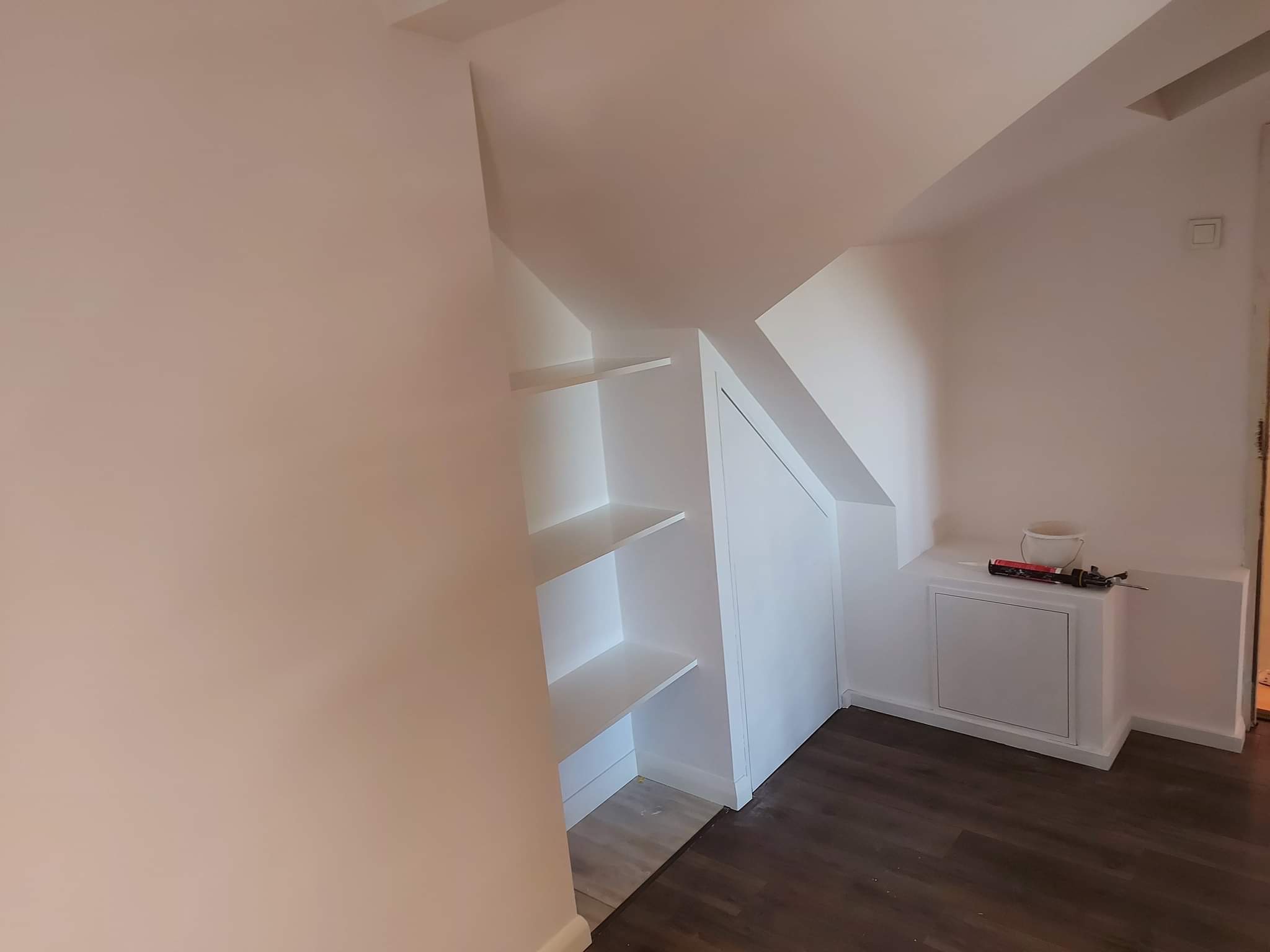 Adaptacja poddasza: zabudowa ze skosem z półkami i szafką, świeżo pomalowane ściany w odcieniach bieli, ciemna podłoga, widoczny silikon i wiadro na szafce.