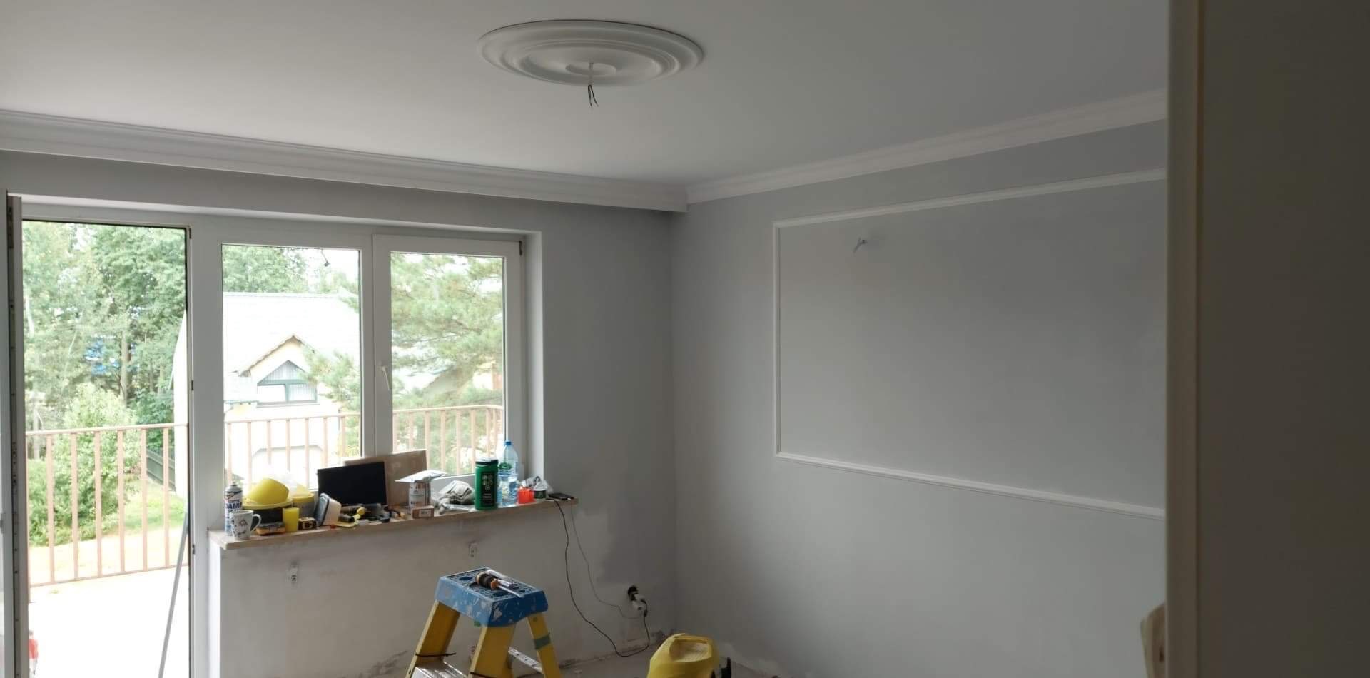 Pokój w trakcie remontu z pomalowanymi na szaro ścianami, listwami dekoracyjnymi pod sufitem i wokół prostokątnego obramowania na ścianie, widoczne narzędzia i akcesoria na parapecie i drabinie.