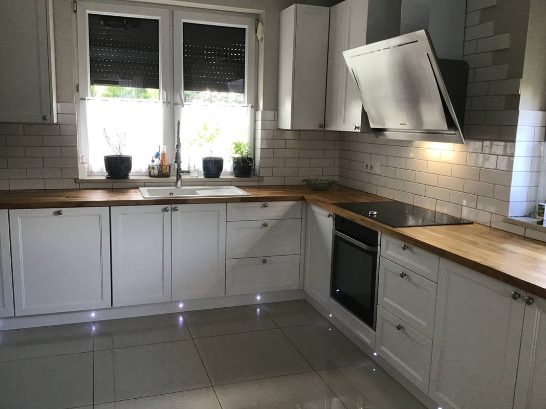 Jasna kuchnia z białymi szafkami, drewnianym blatem i podświetleniem LED pod szafkami, widok na okno z roletami.