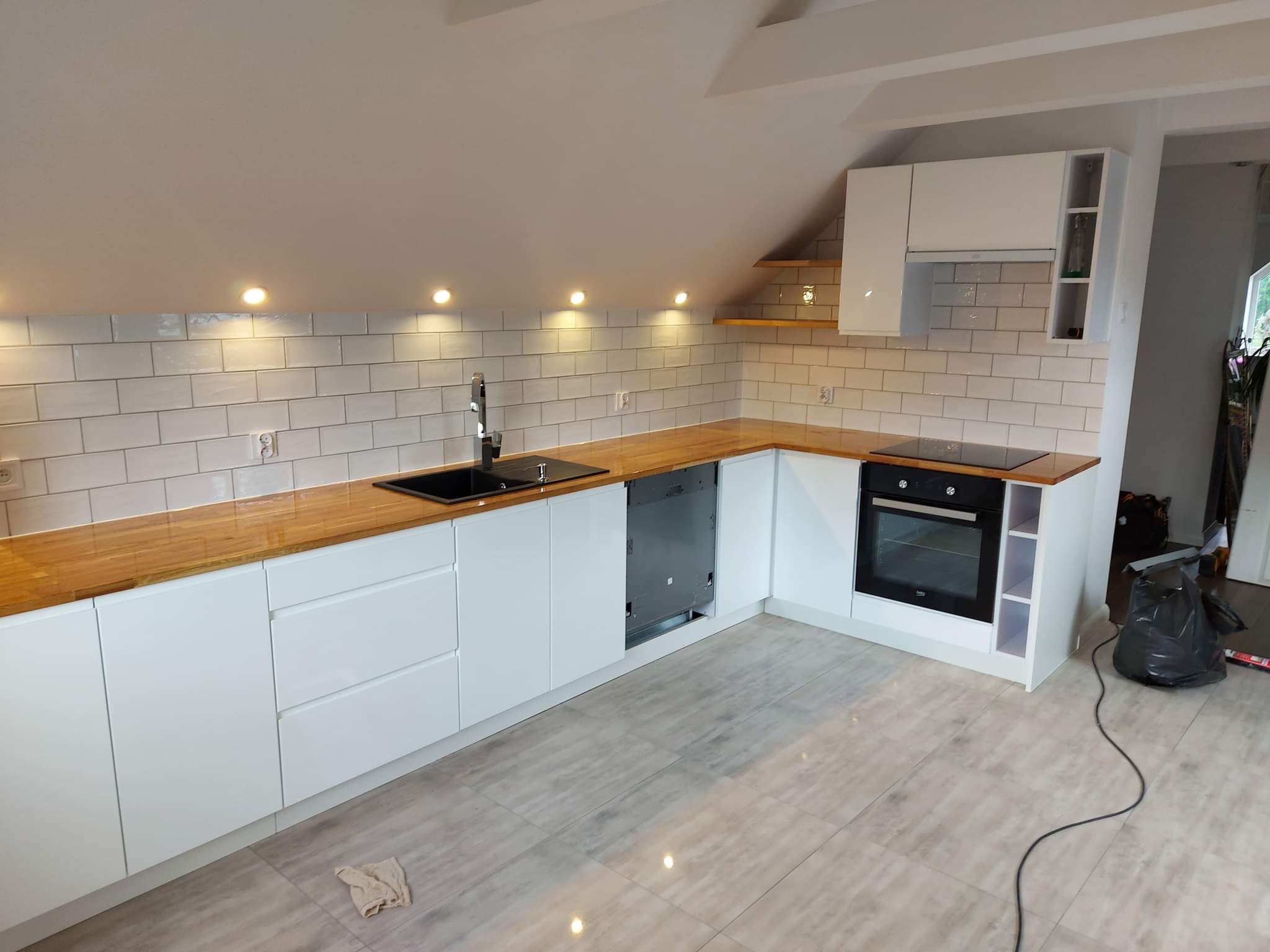 Nowoczesna kuchnia w skosach, białe szafki z drewnianym blatem, płytki imitujące cegłę, oświetlenie punktowe podszafkowe, płyta indukcyjna i piekarnik w zabudowie, szara podłoga z płytek.