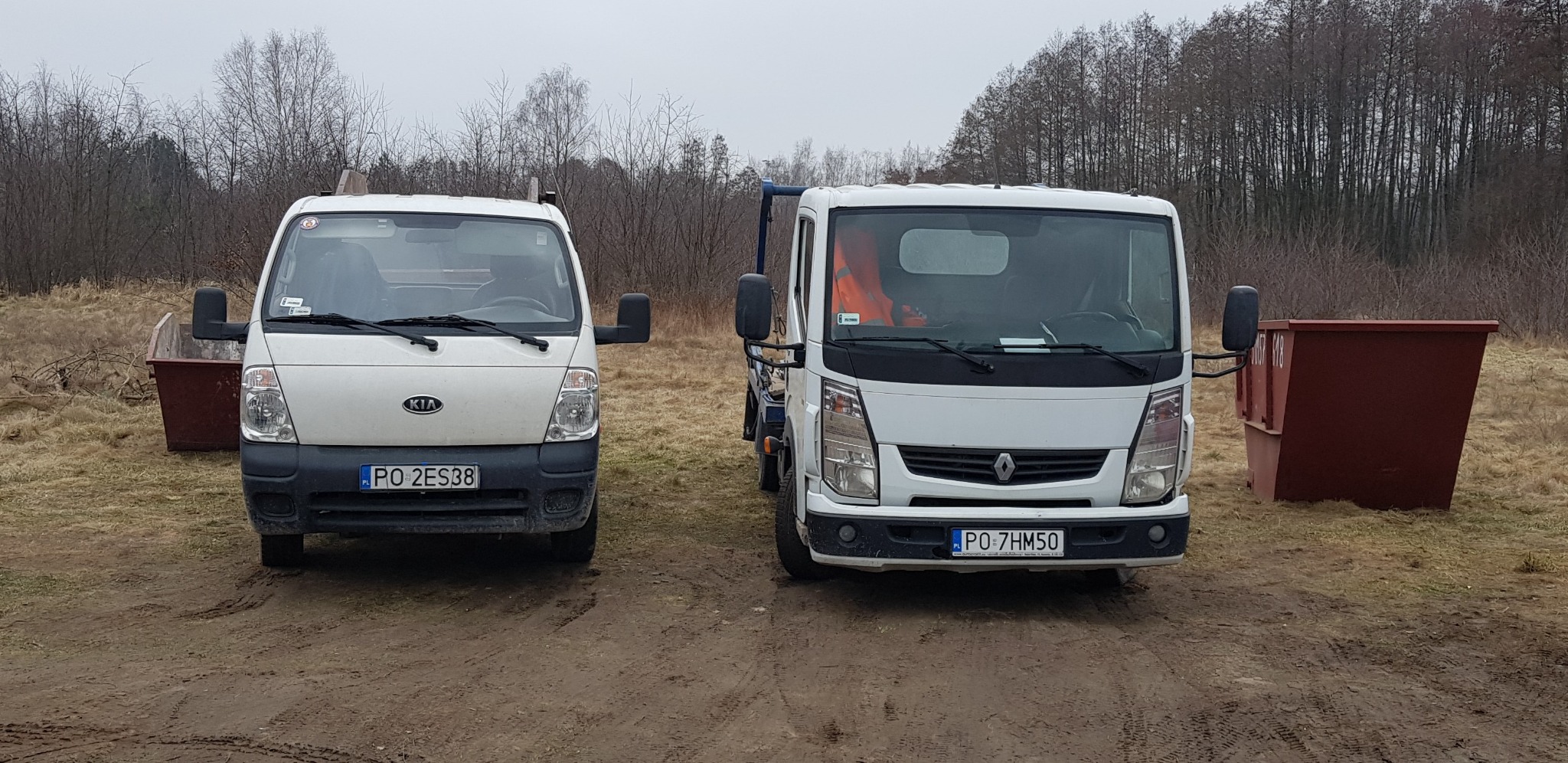 Dwa białe samochody dostawcze (Kia i Renault) stojące na ziemi obok kontenerów na gruz w plenerze.