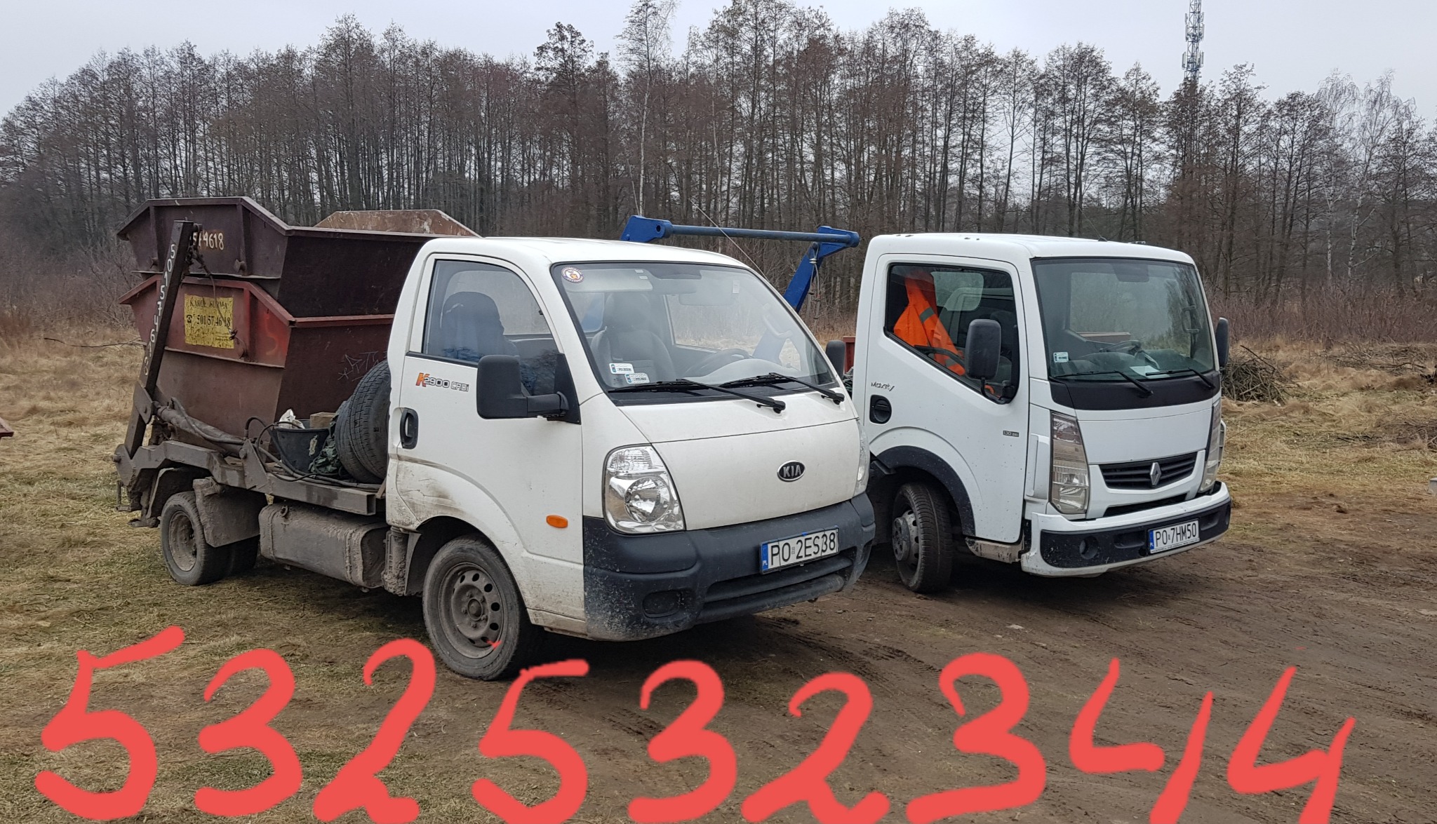 Dwa białe samochody ciężarowe, jeden z kontenerem na gruz, stoją na ziemi w pobliżu drzew. Kontener jest częściowo wypełniony. Widoczna tablica rejestracyjna pojazdu.