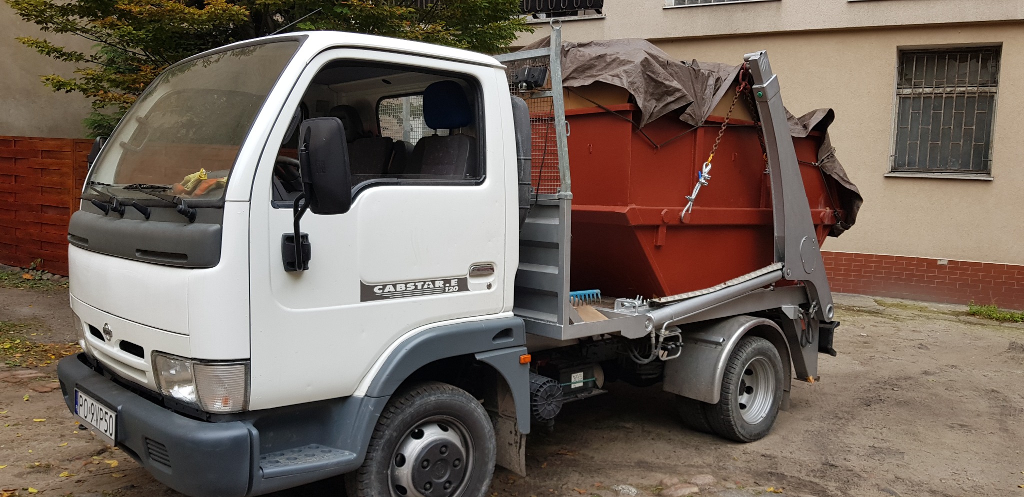 Biała ciężarówka Nissan Cabstar z czerwonym, częściowo przykrytym plandeką kontenerem na gruz na platformie, zaparkowana na podwórku z widoczną elewacją budynku i fragmentem zieleni.