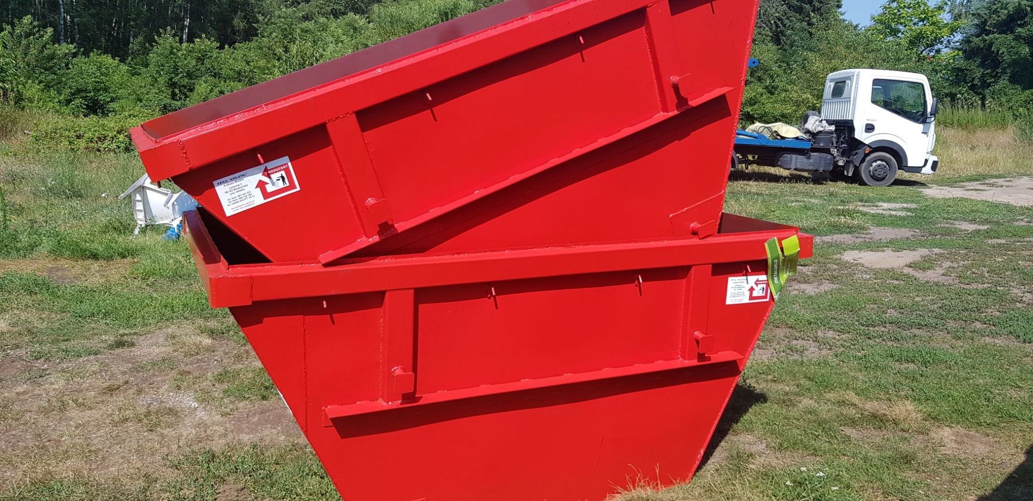 Dwa czerwone, puste kontenery na gruz ustawione jeden na drugim, z widoczną naklejką producenta i białą ciężarówką w tle na trawiastym terenie.