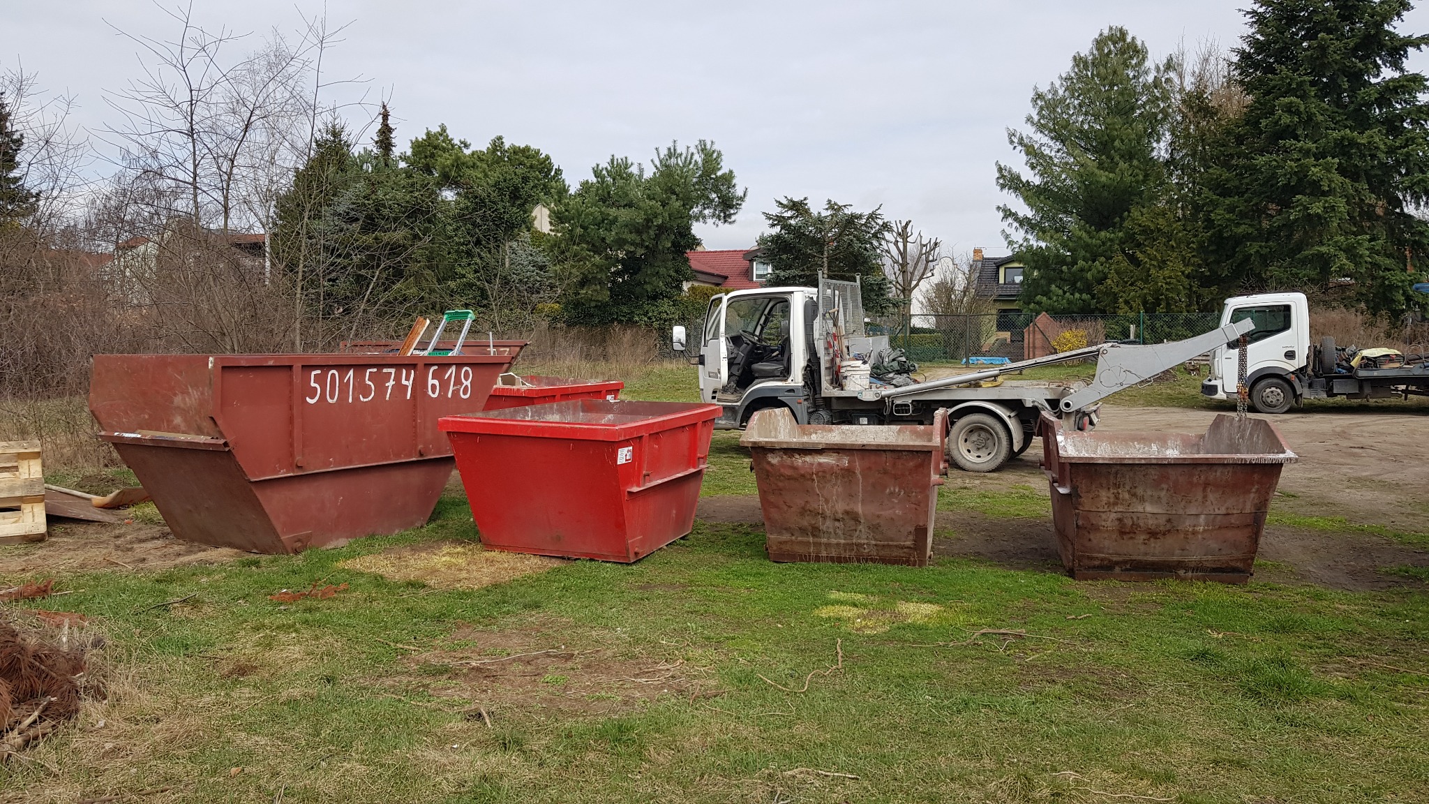 Kontenery na gruz różnych rozmiarów i kolorów (czerwony i brązowy) oraz samochód ciężarowy z HDS do ich transportu, na trawiastym terenie w Poznaniu.
