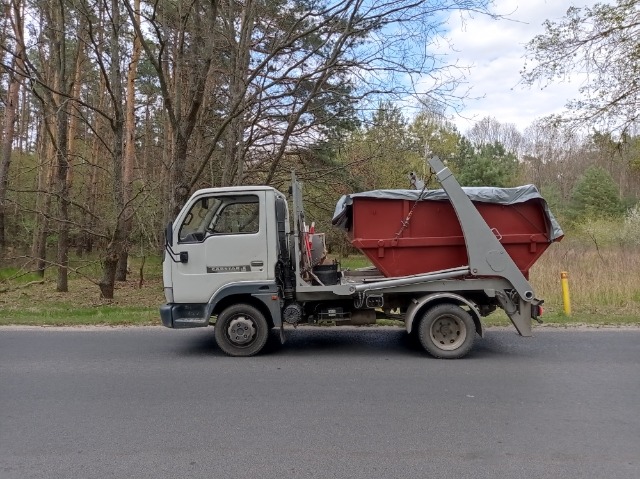 Biała ciężarówka z czerwonym kontenerem na gruz, zabezpieczonym szarą plandeką, stojąca na poboczu drogi w otoczeniu drzew.
