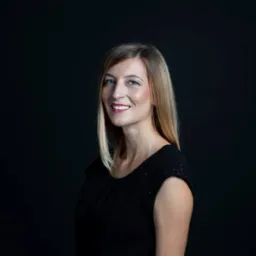 Portret uśmiechniętej kobiety z blond włosami w czarnej sukience na ciemnym tle.