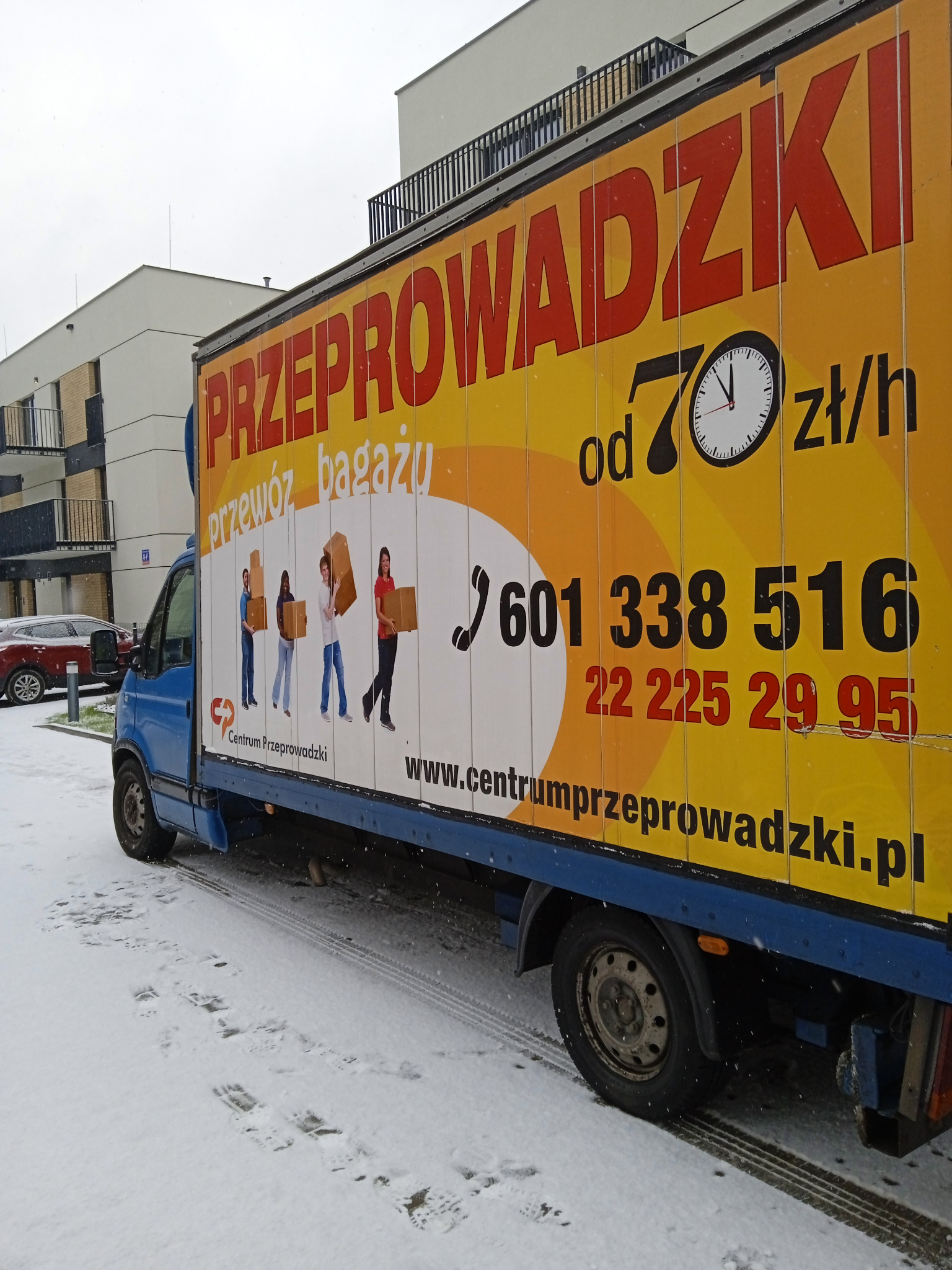 Niebieski samochód dostawczy z reklamą firmy oferującej usługi transportowe, zaparkowany na śniegu przed nowoczesnym budynkiem mieszkalnym. Na żółtym tle nadrukowane są duże czerwone litery z nazwą...