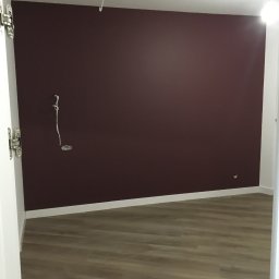 HomePlus - wykończenia pod klucz Mateusz Kossowski - Pomalowane na burgundowo ściany w pustym pokoju z panelami podłogowymi, widoczne wystające kable elektryczne i białe listwy przypodłogowe, ujęcie zza otwartych drzwi.