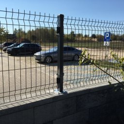 HomePlus - wykończenia pod klucz Mateusz Kossowski - Nowoczesne ogrodzenie panelowe z metalowej siatki na betonowej podmurówce, widok na parking z zaparkowanymi samochodami i znak dla niepełnosprawnych, w tle drzewa i plac budowy z dźwigiem.