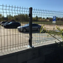Nowoczesne ogrodzenie panelowe z metalowej siatki na betonowej podmurówce, widok na parking z zaparkowanymi samochodami i znak dla niepełnosprawnych, w tle drzewa i plac budowy z dźwigiem.