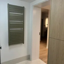 Nowoczesna łazienka z grzejnikiem drabinkowym w kolorze oliwkowym, jasnymi płytkami podłogowymi imitującymi marmur i widokiem na dalszą część mieszkania przez otwarte drzwi.