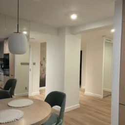 Jasne wnętrze mieszkania z okrągłym drewnianym stołem, zielonymi krzesłami i podkładkami, widok na korytarz z tapetą w kwiaty i lustrzanymi drzwiami.