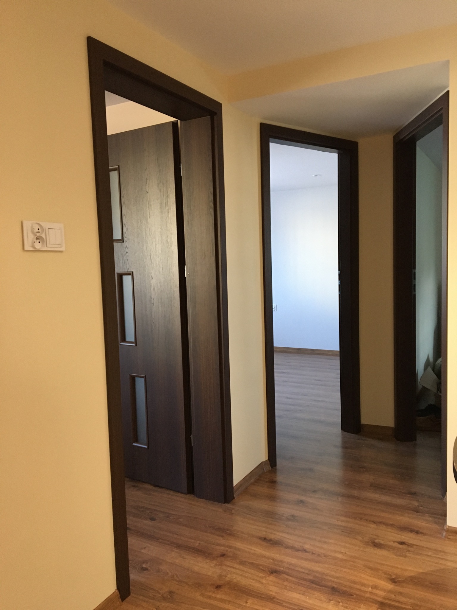 Wnętrze mieszkania z trzema parami otwartych drzwi w ciemnych, drewnianych ramach; podłoga wyłożona panelami imitującymi drewno, ściany w odcieniach beżu.