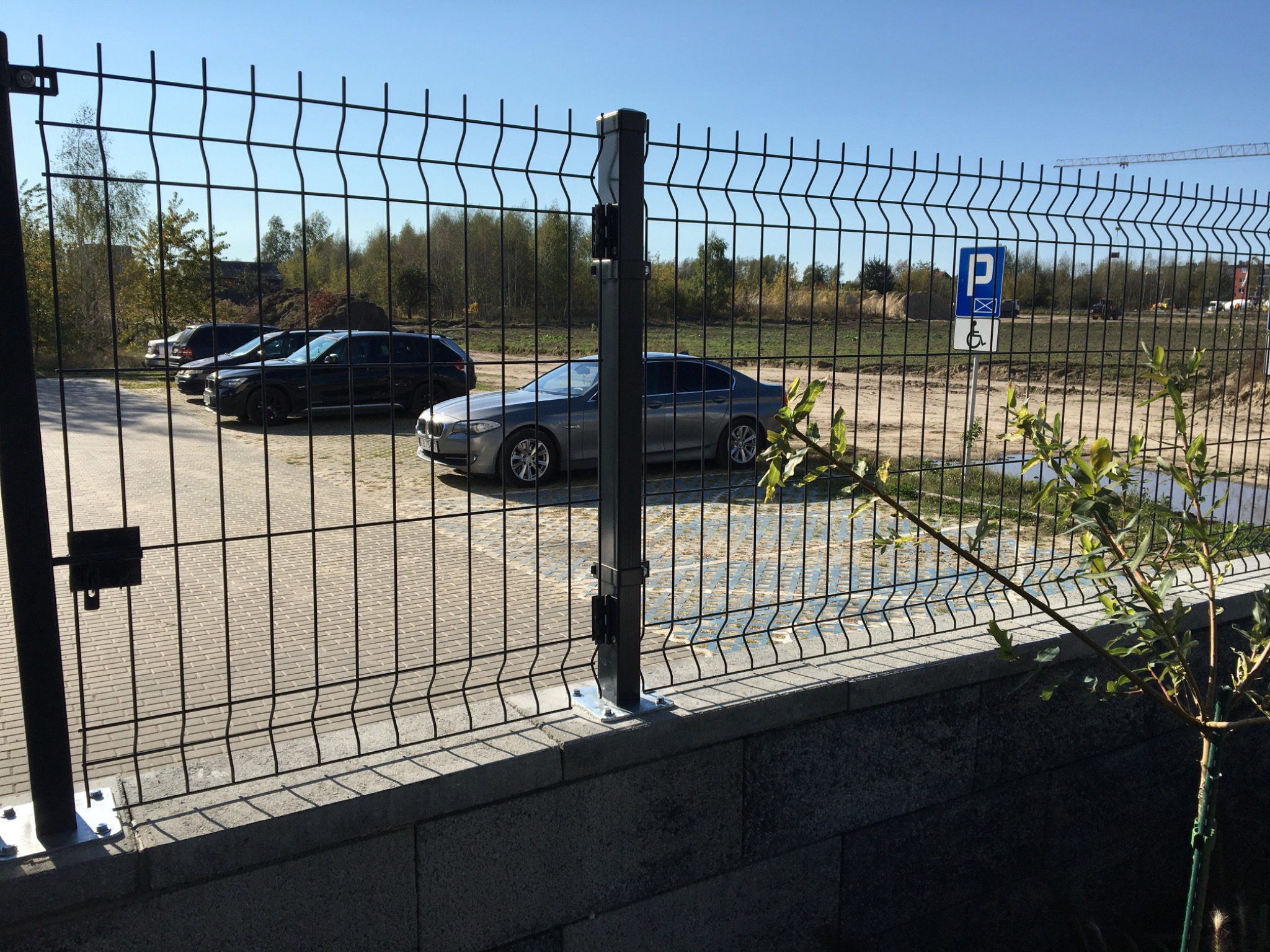 Nowoczesne ogrodzenie panelowe z metalowej siatki na betonowej podmurówce, widok na parking z zaparkowanymi samochodami i znak dla niepełnosprawnych, w tle drzewa i plac budowy z dźwigiem.
