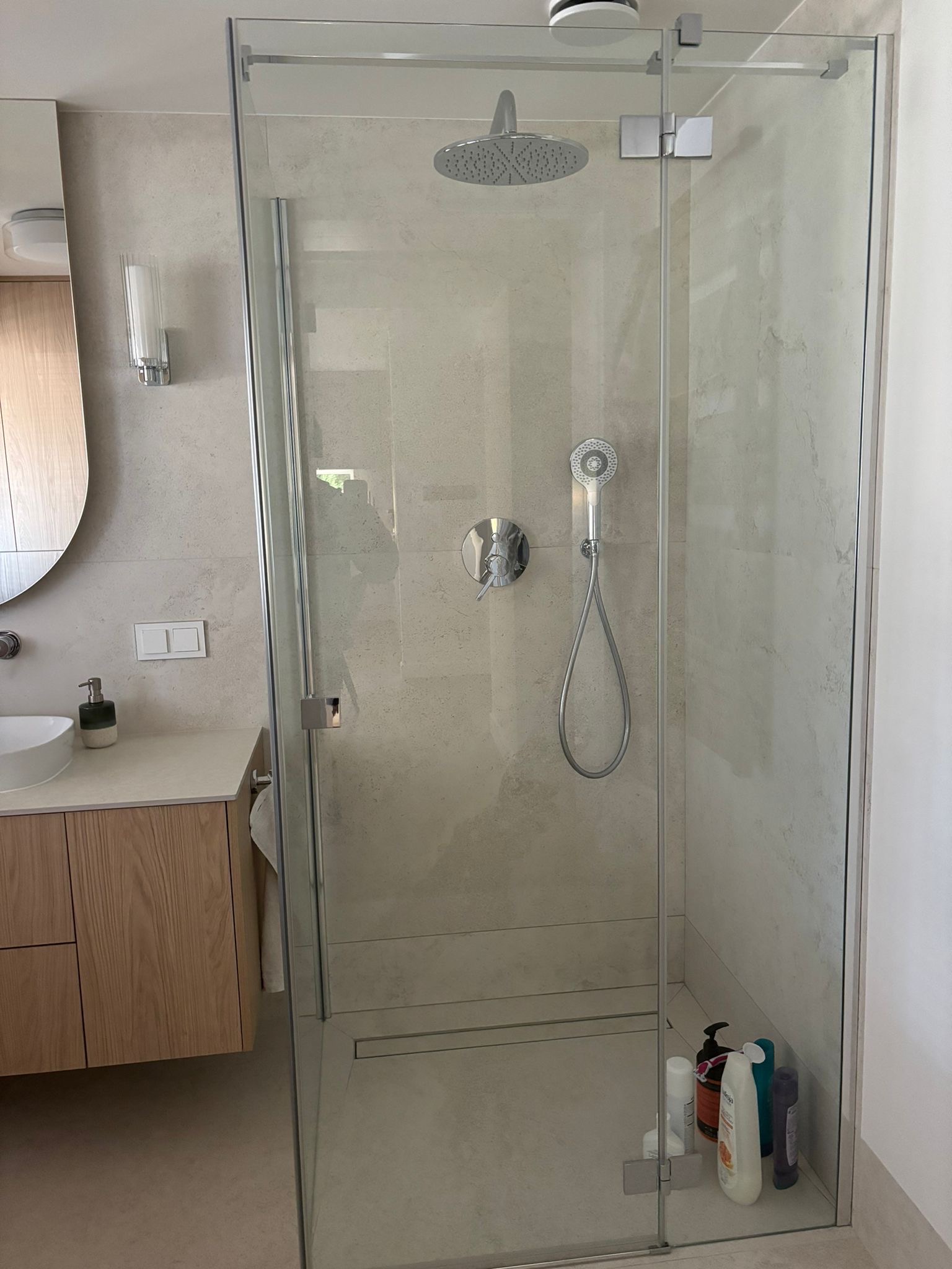 Nowoczesna kabina prysznicowa ze szklanymi drzwiami, deszczownicą i armaturą w kolorze chromu, widoczne kosmetyki wewnątrz kabiny, beżowe płytki na ścianach i drewniana szafka umywalkowa.