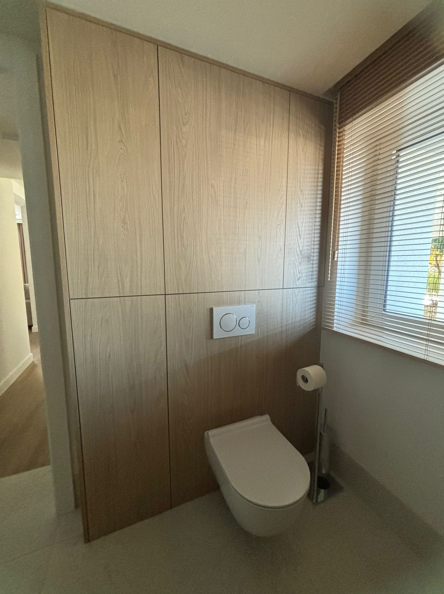 Nowoczesna łazienka z zabudową meblową z jasnego drewna, wisząca toaleta z chromowanym przyciskiem spłukiwania i rolety okienne w tle.