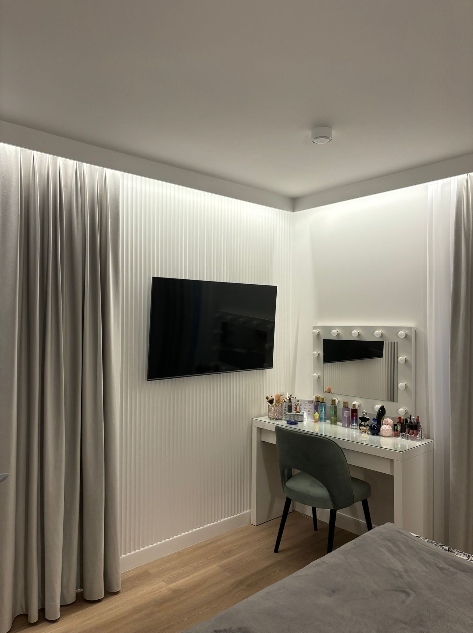 Nowoczesna sypialnia z toaletką z lustrem podświetlanym żarówkami, telewizorem na ścianie z dekoracyjnymi panelami pionowymi i zasłonami w odcieniach szarości.