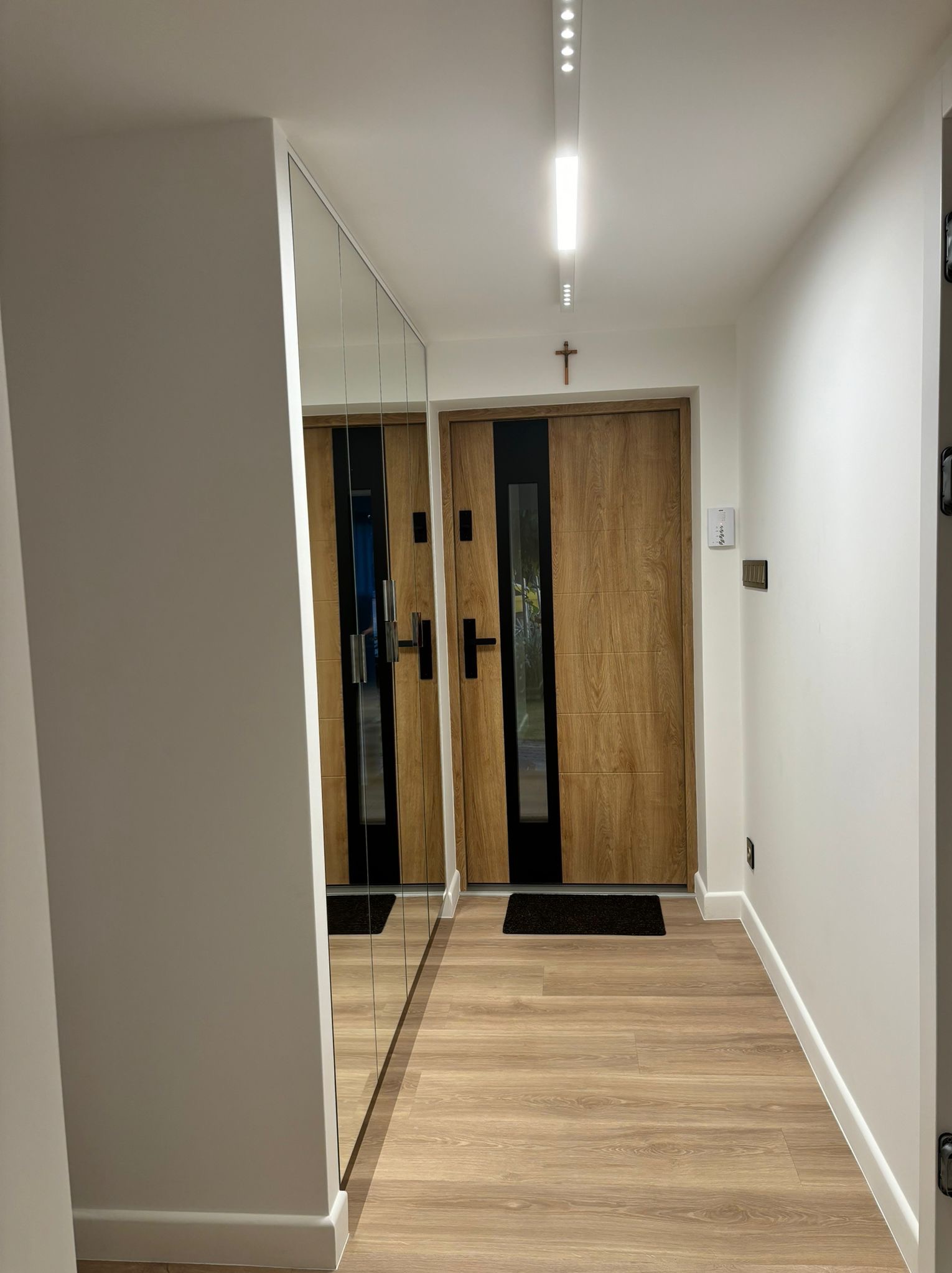 Nowoczesny przedpokój z drewnianą podłogą, lustrzaną szafą wnękową i dębowymi drzwiami z czarnymi wstawkami, oświetlony ledowym panelem sufitowym.