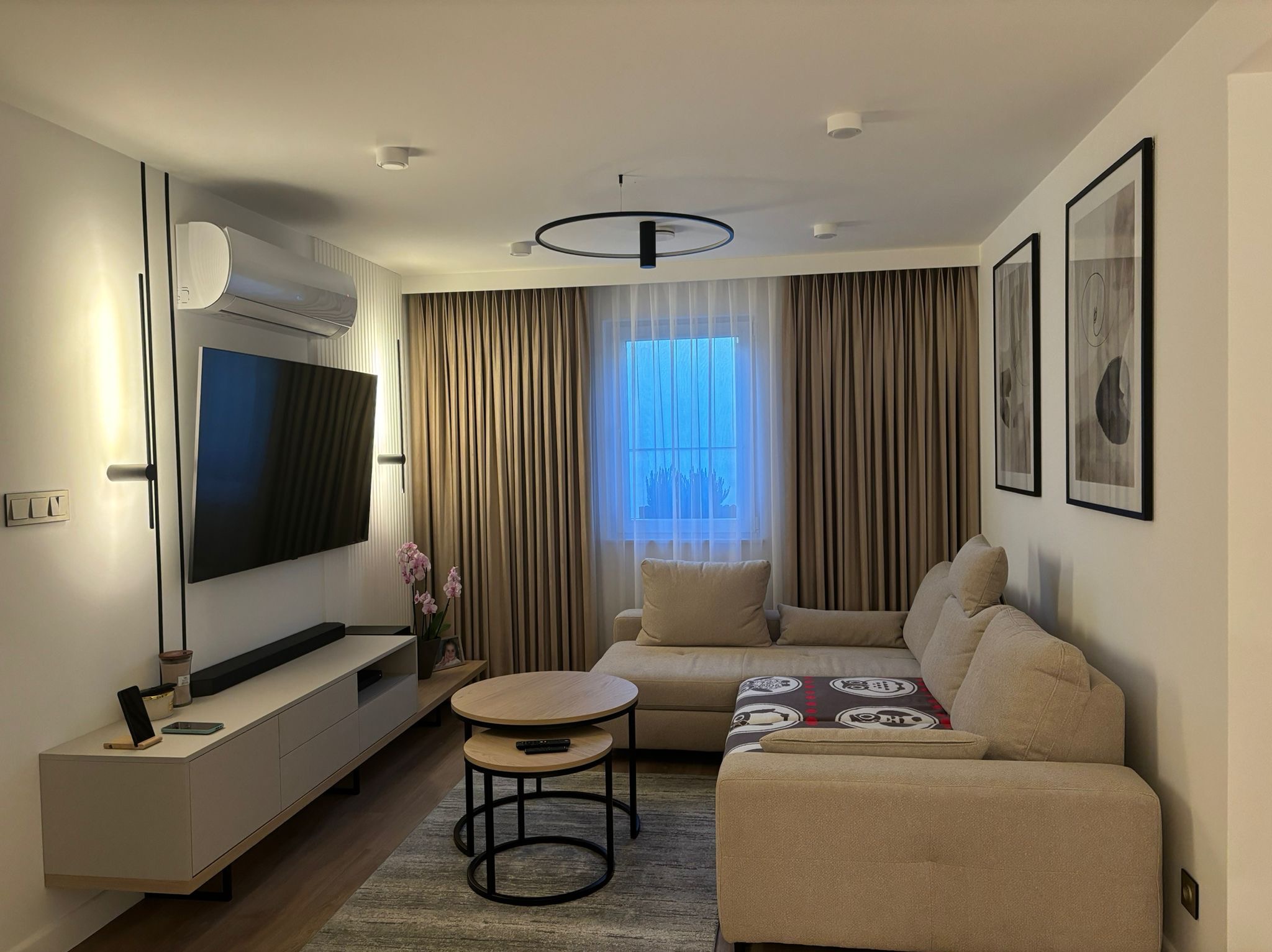 Nowoczesny salon z narożną sofą w beżowych odcieniach, stolikiem kawowym z dwoma blatami, telewizorem na ścianie i klimatyzatorem, oświetlony okrągłą lampą sufitową.