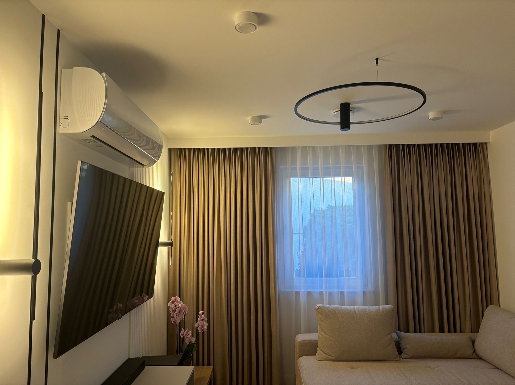 Nowoczesne wnętrze salonu z klimatyzacją, telewizorem na ścianie, minimalistyczną lampą sufitową i zasłonami w odcieniach beżu.