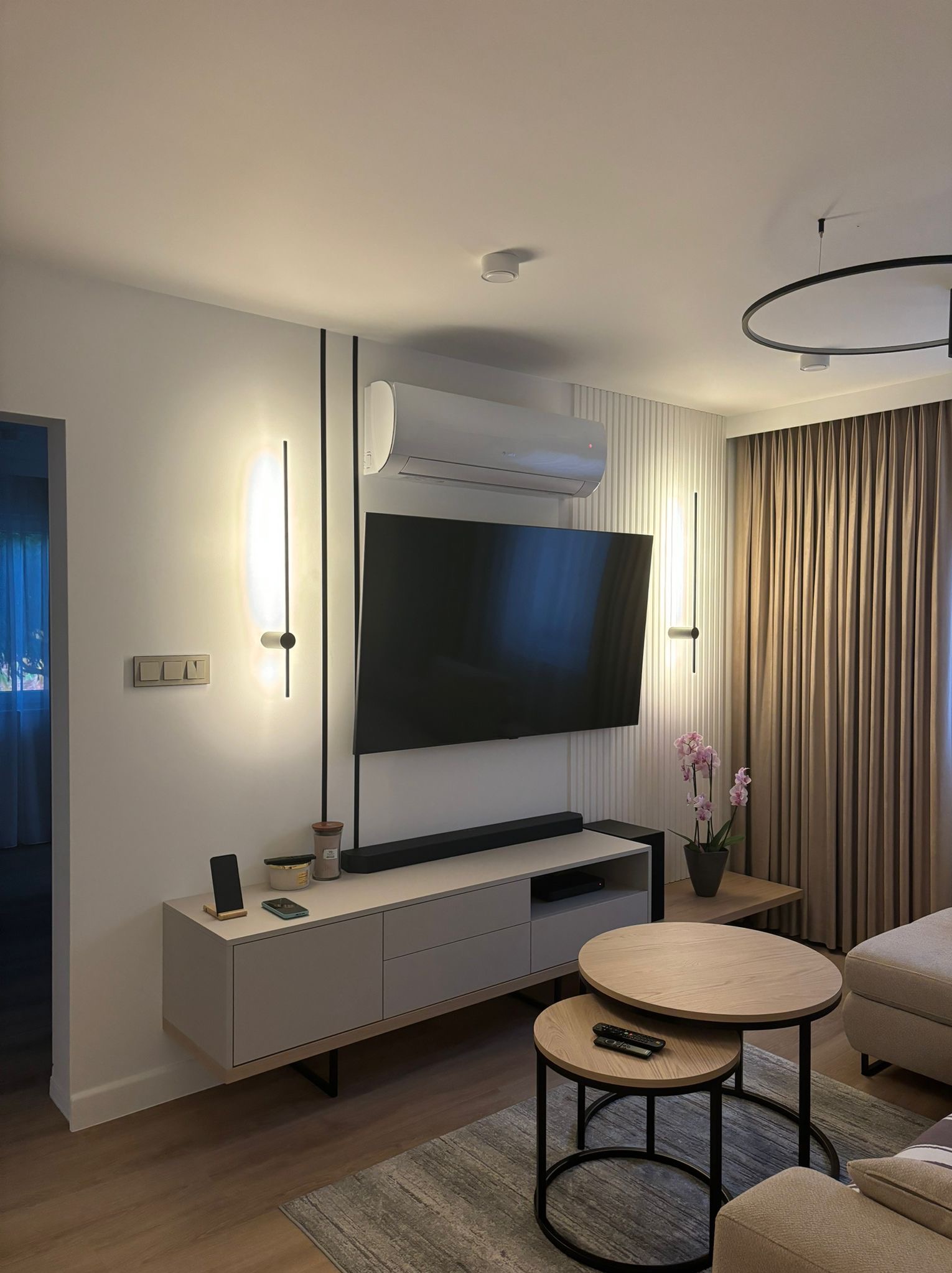 Nowoczesny salon z beżową szafką RTV, wiszącym telewizorem, klimatyzatorem, okrągłymi stolikami kawowymi i dekoracyjnymi lampami ściennymi.