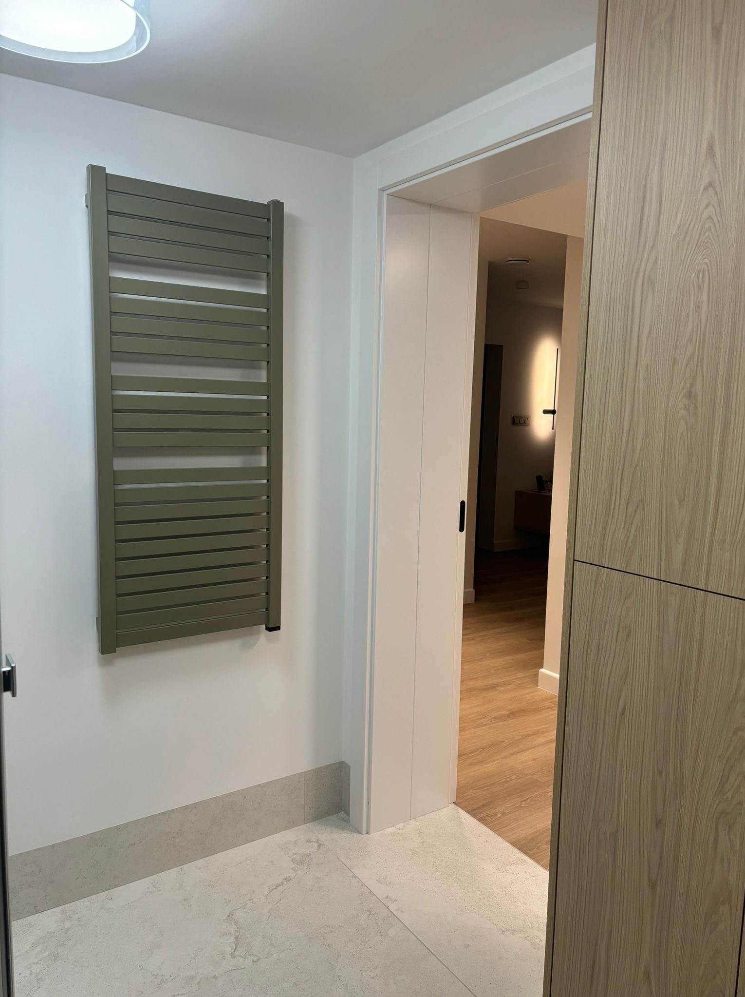 Nowoczesna łazienka z grzejnikiem drabinkowym w kolorze oliwkowym, jasnymi płytkami podłogowymi imitującymi marmur i widokiem na dalszą część mieszkania przez otwarte drzwi.