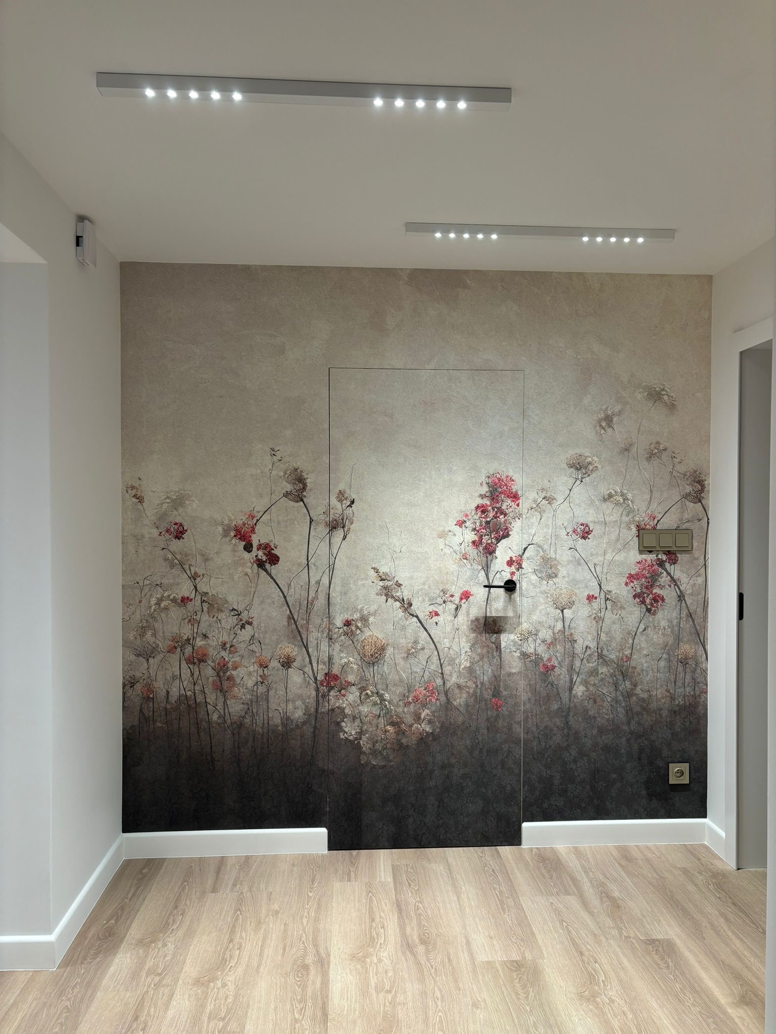 Nowoczesny przedpokój z fototapetą w motywy kwiatowe, ukryte drzwi, oświetlenie sufitowe LED i panele podłogowe imitujące drewno.