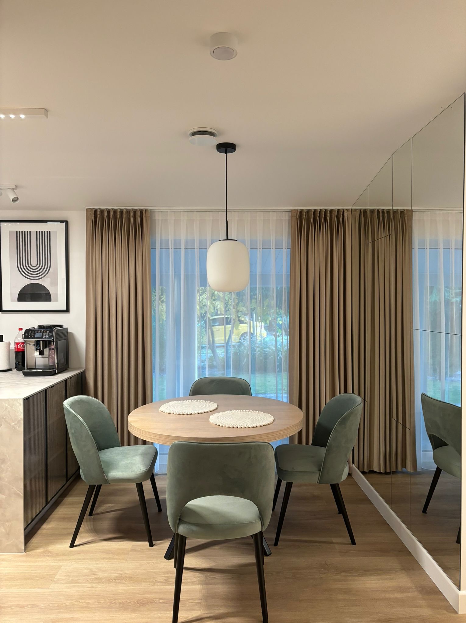 Jasne wnętrze jadalni z okrągłym drewnianym stołem, czterema welurowymi krzesłami w odcieniach zieleni, minimalistyczną lampą wiszącą i lustrzaną ścianą, uzupełnione zasłonami w kolorze beżowym...