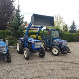 AGRO-PARK - Trzy niebieskie traktory Iseki na żwirowym podłożu, z różnymi przystawkami, w tle zieleń drzew i krzewów. Jeden z traktorów ma zamontowaną łyżkę.