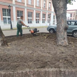 AGRO-PARK - Mężczyzna w zielonym kombinezonie używa glebogryzarki do przygotowania ziemi pod nasadzenia w miejskiej rabacie z obrzeżem, na tle budynku z czerwonej cegły.